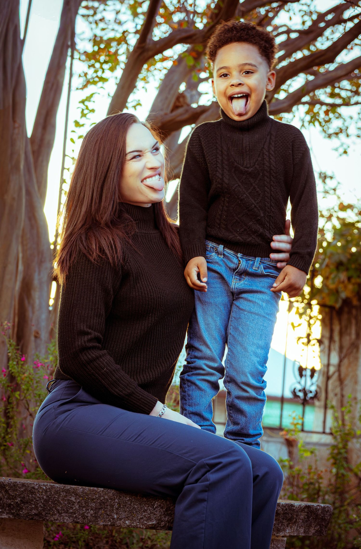 A couple sits together on a stone wall at sunset wearing black sweaters and jeans in a romantic outdoor setting.