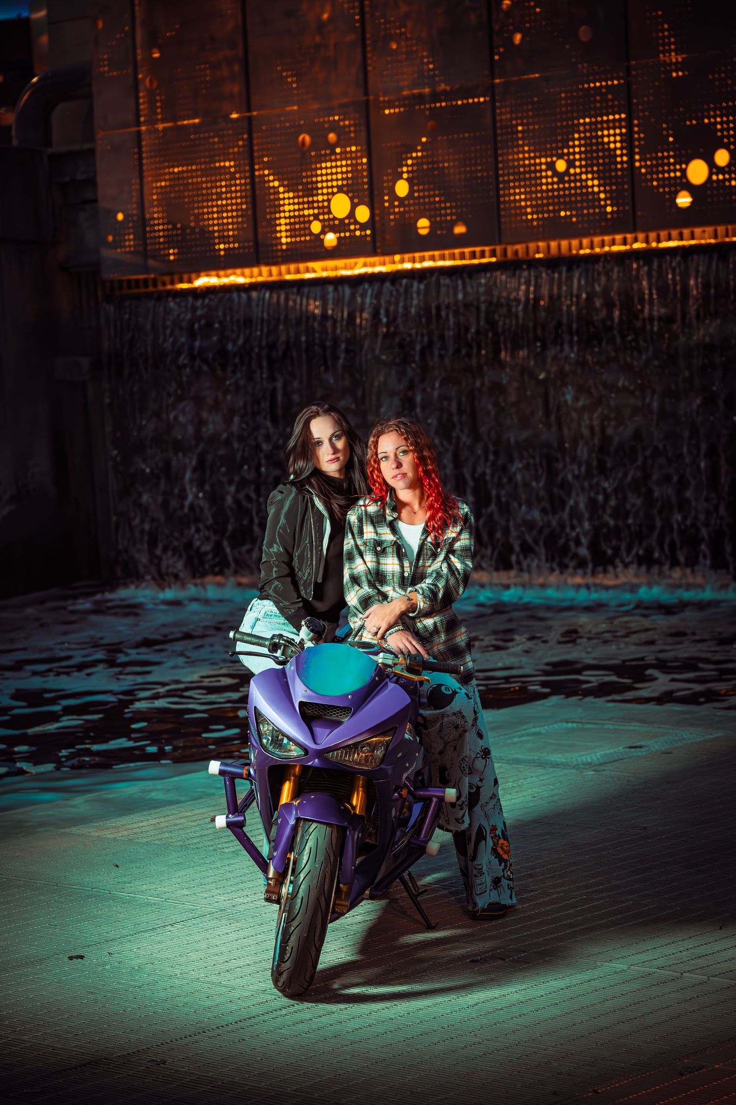 Two riders pose on a motorcycle at night with dramatic blue and orange lighting creating a cinematic atmosphere.