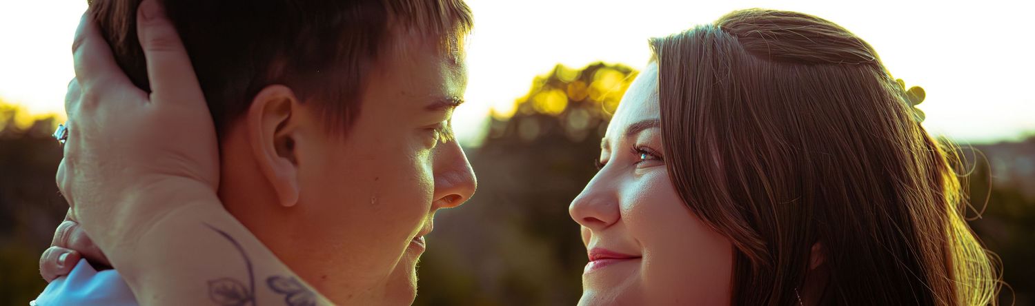 A couple gazes at each other romantically outdoors during golden hour, the man with tattoos and the woman smiling softly.