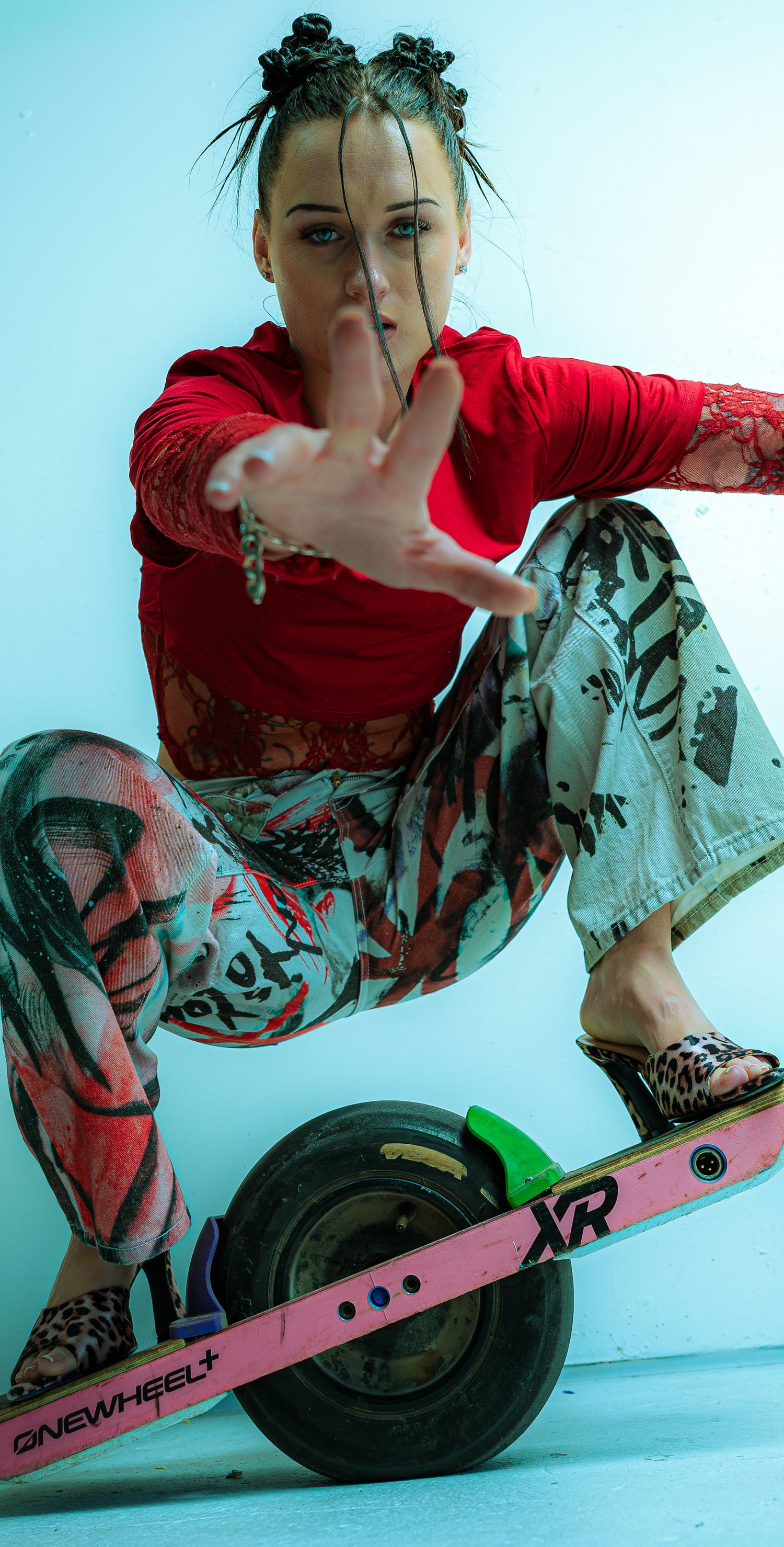 Woman in red top and graphic pants balancing on a Onewheel XR electric skateboard while reaching toward camera.