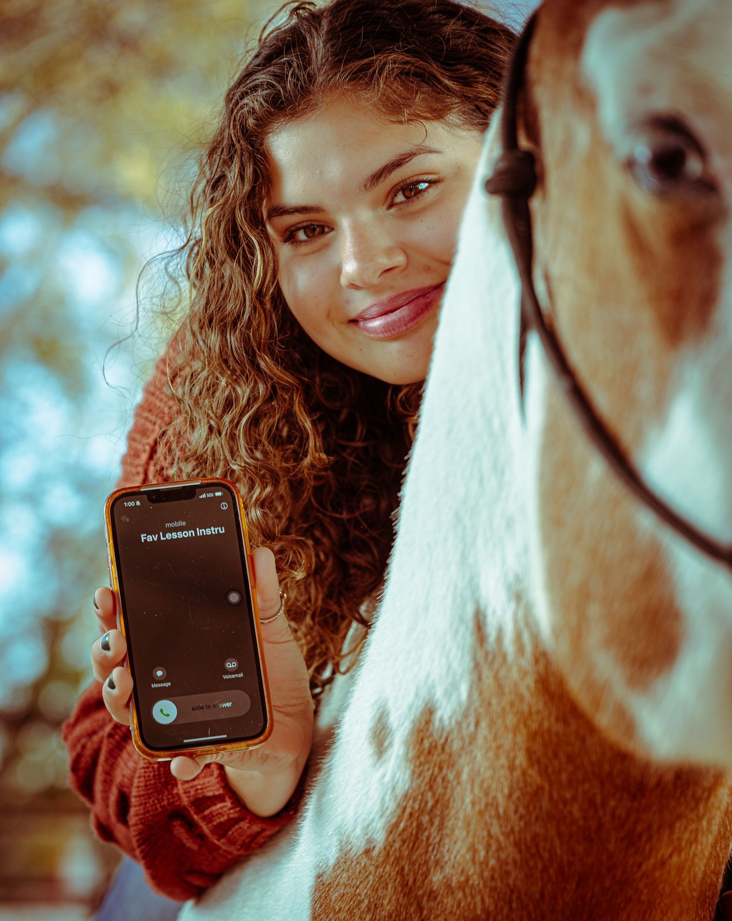 Person with curly hair smiling while holding a mobile phone displaying a dark interface in warm lighting.