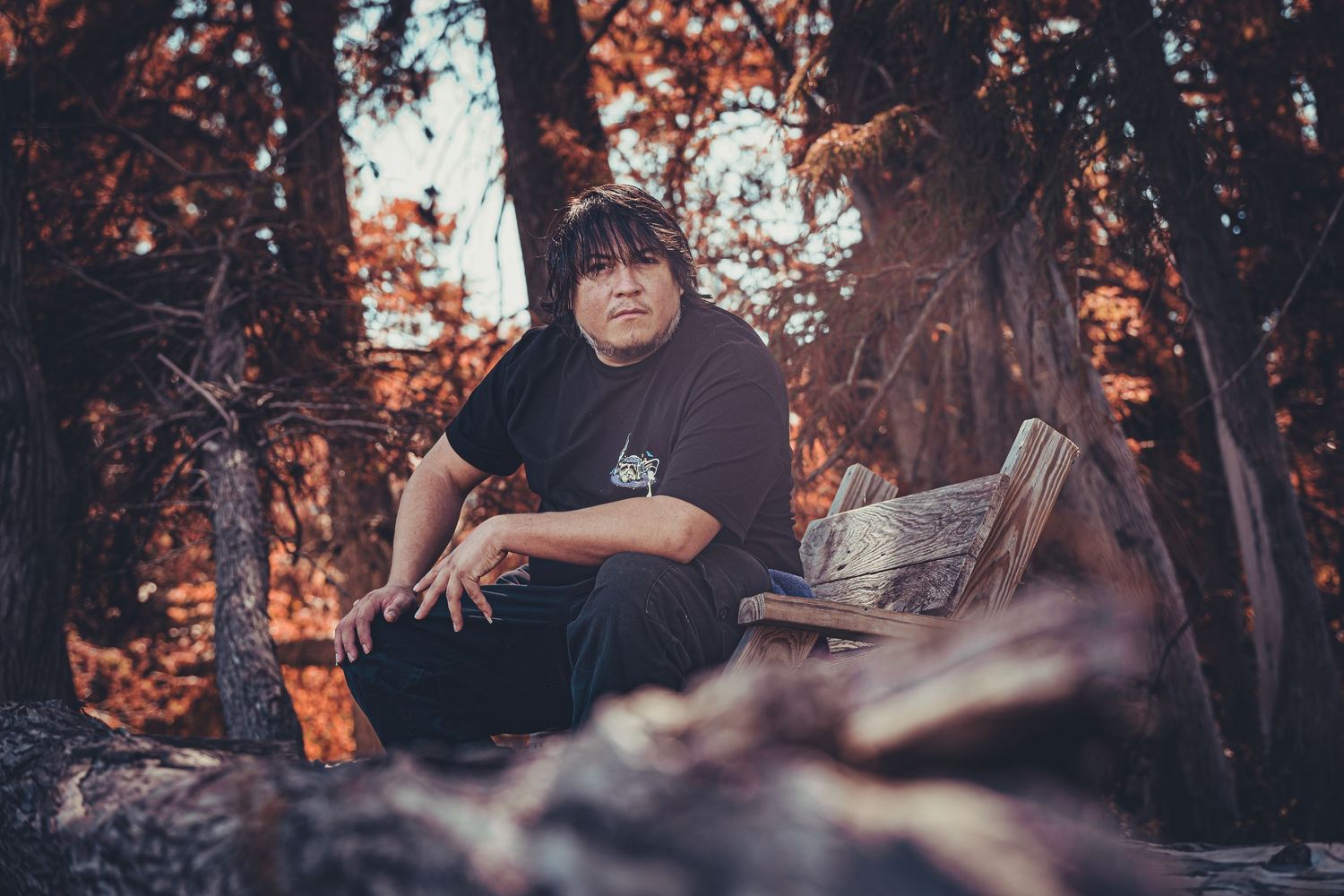 Person in black clothing sitting casually in a wooded area during autumn.