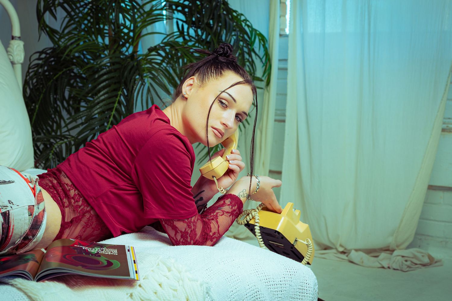 Woman in red lace outfit lying on bed talking on yellow retro phone surrounded by plants and white curtains.