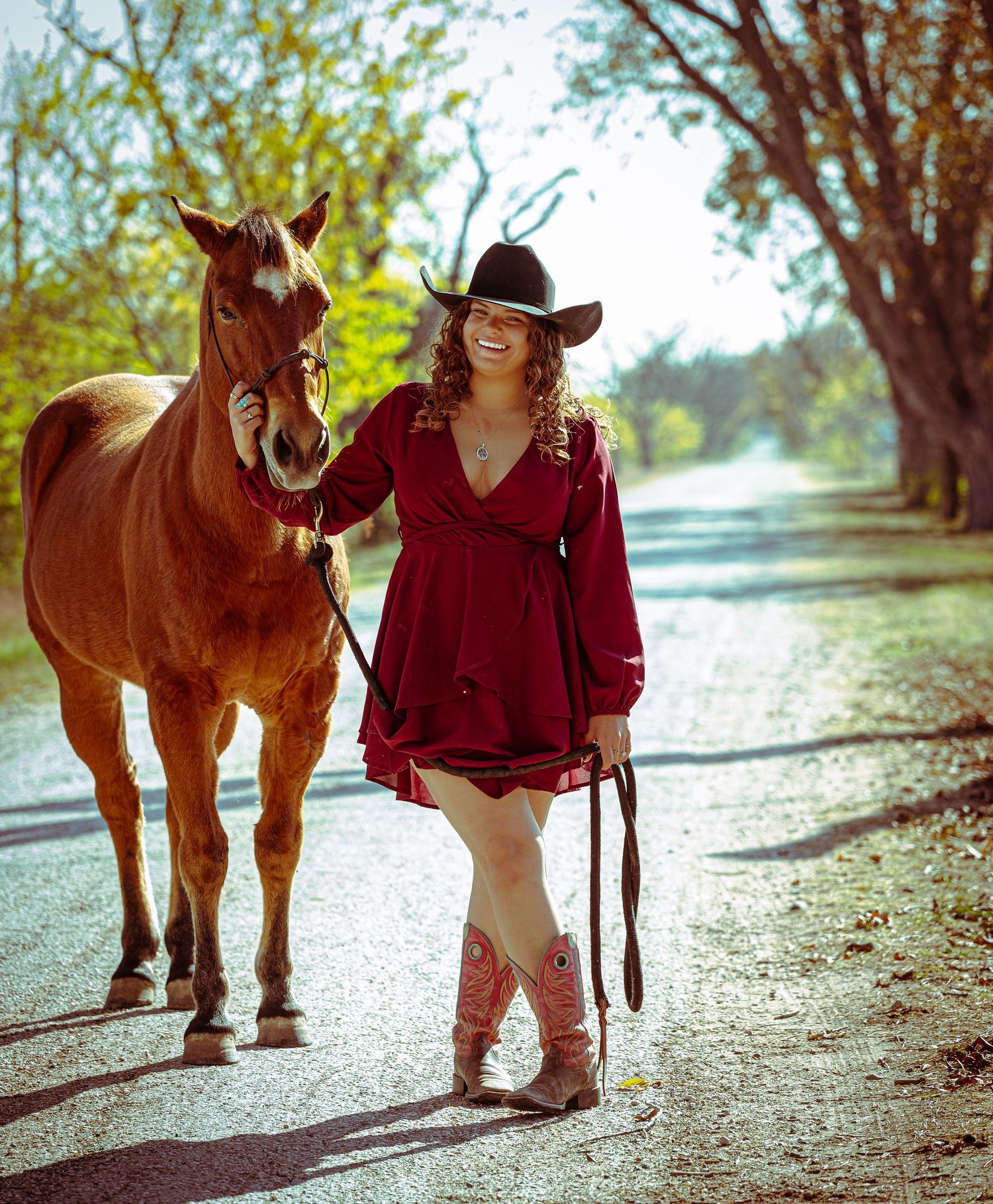 A person in a burgundy sweater and cowboy hat walks with a chestnut horse along a tree-lined path.