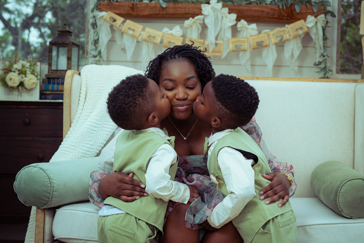 A mother smiles joyfully as her two young sons in green suits lean in to kiss her cheeks simultaneously.