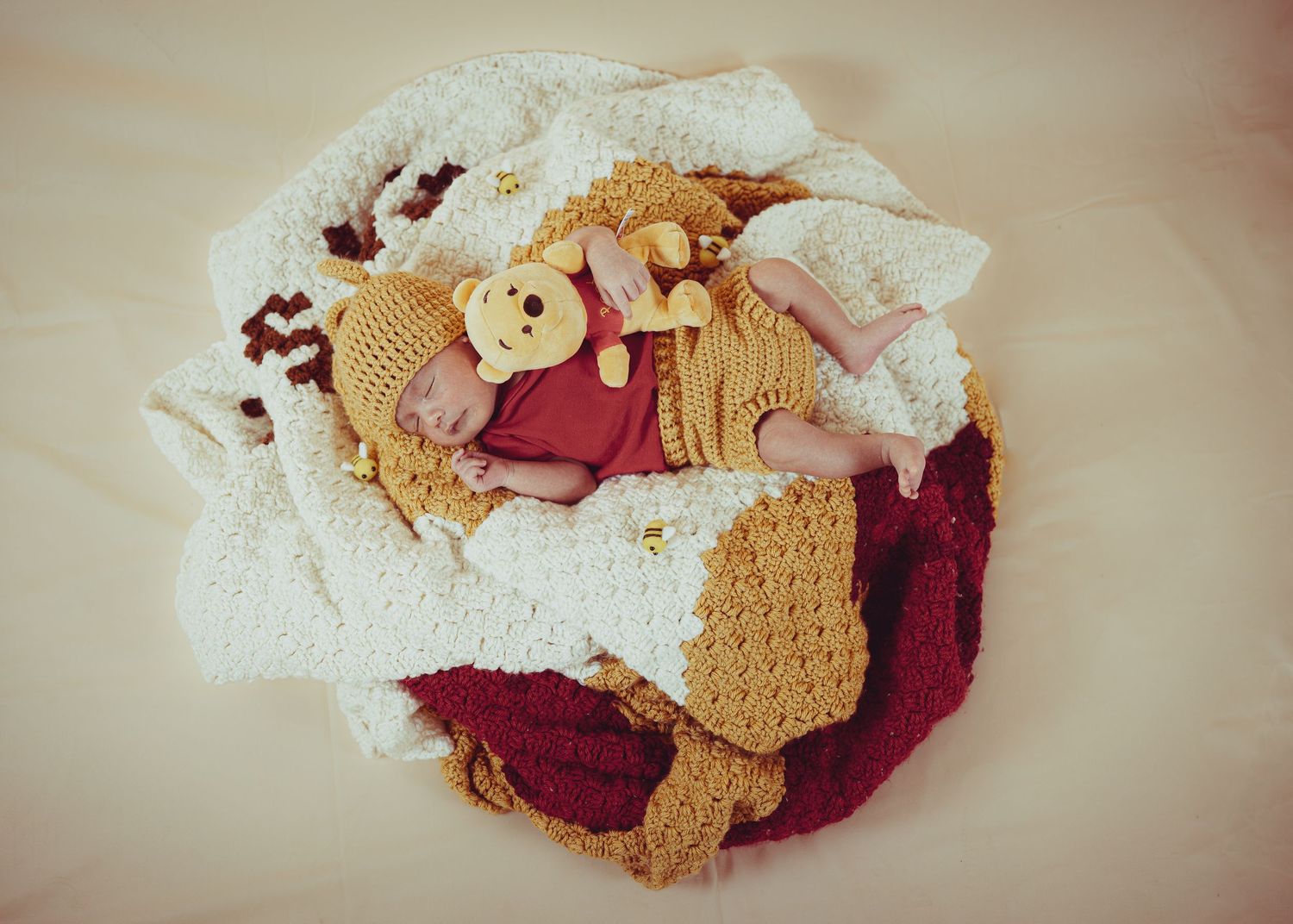 Newborn baby sleeping in a nest of colorful crocheted blankets wearing a Winnie the Pooh hat and red outfit.