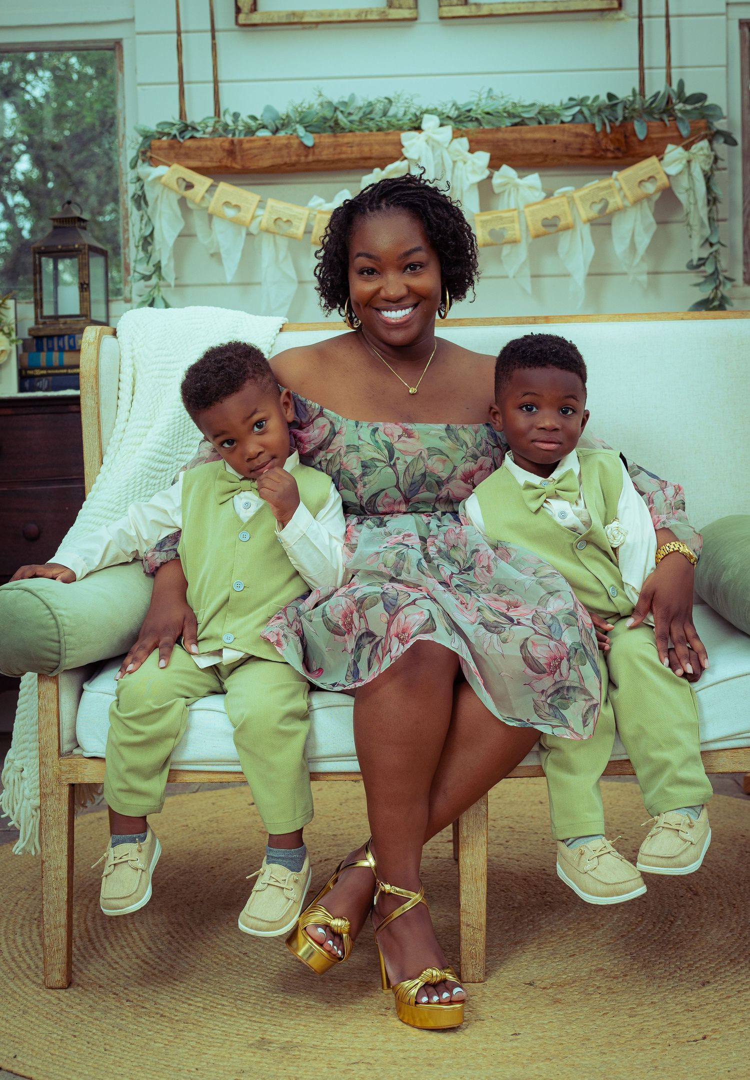 A smiling woman in a floral dress sits with two young boys in matching green suits on a white bench.