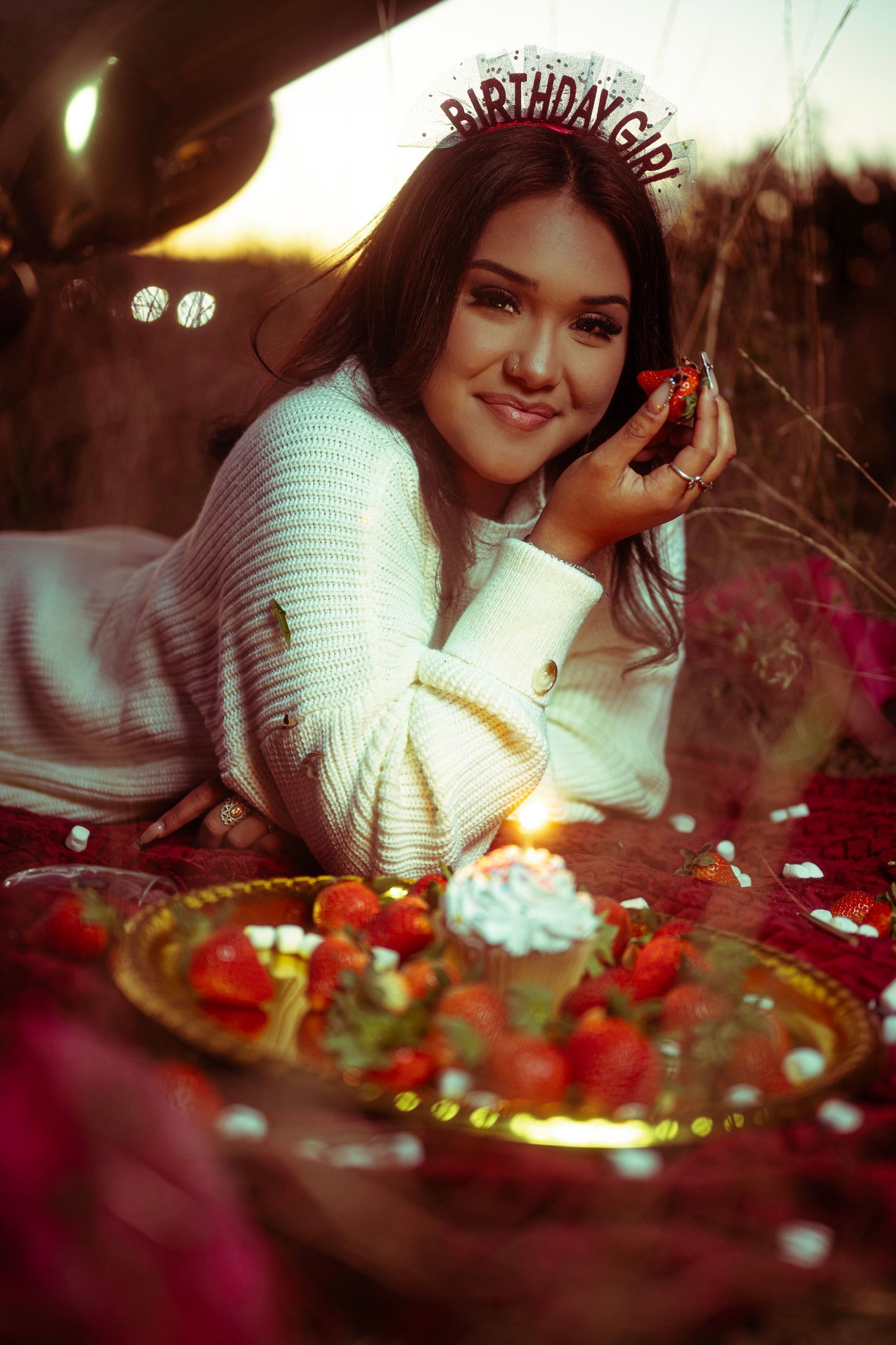 A person in white leans over a festive birthday cake with candles and decorations in a warm, dimly lit setting.