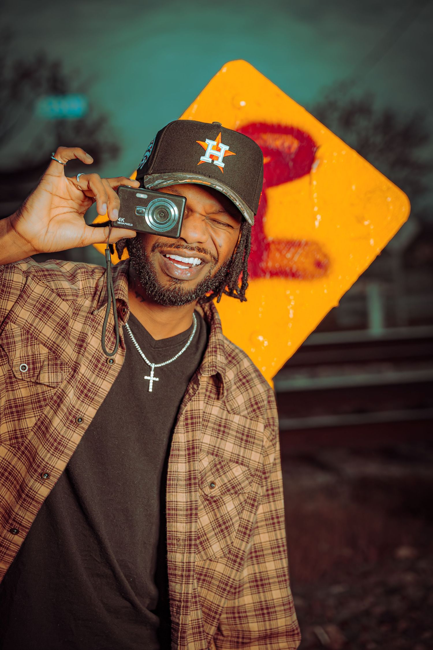 Person in plaid shirt and dark cap laughs while holding a camera against orange background.