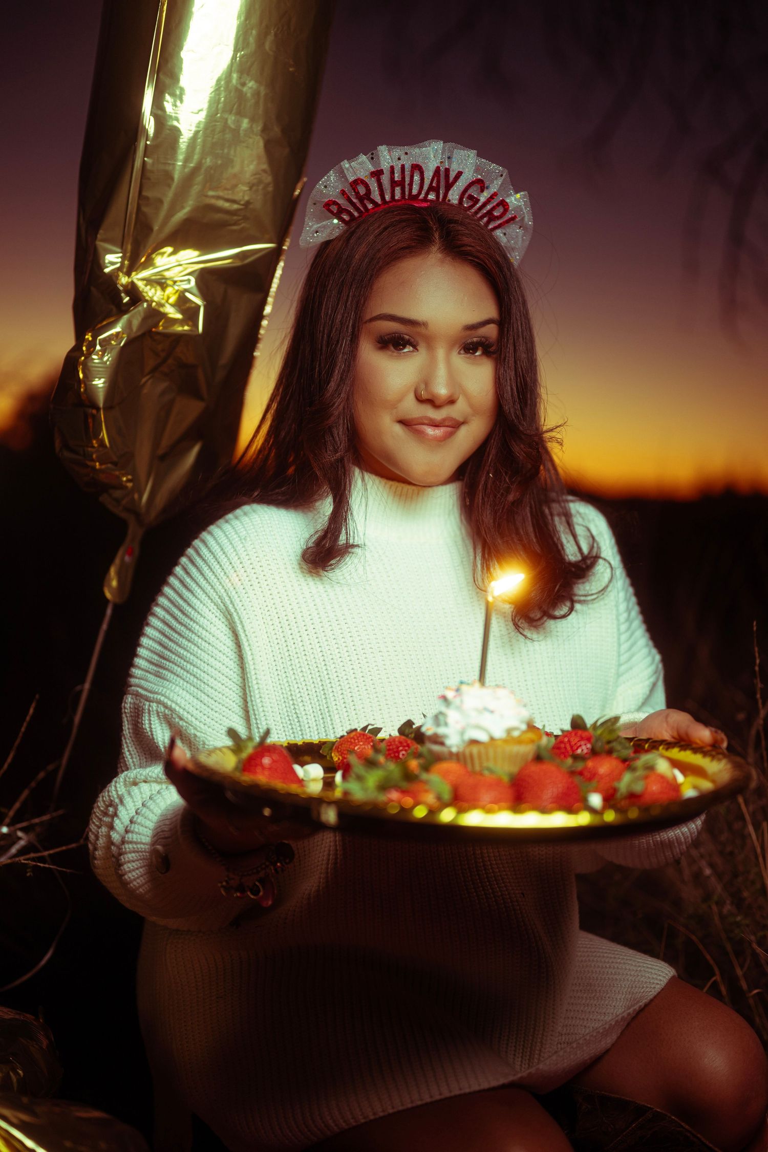 A person in a white sweater sits at a candlelit table with a birthday cake glowing in the warm sunset light.