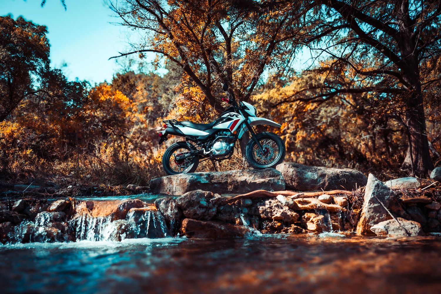 Adventure motorcycle parked near scenic waterfall with autumn foliage and rocky terrain in dramatic lighting.