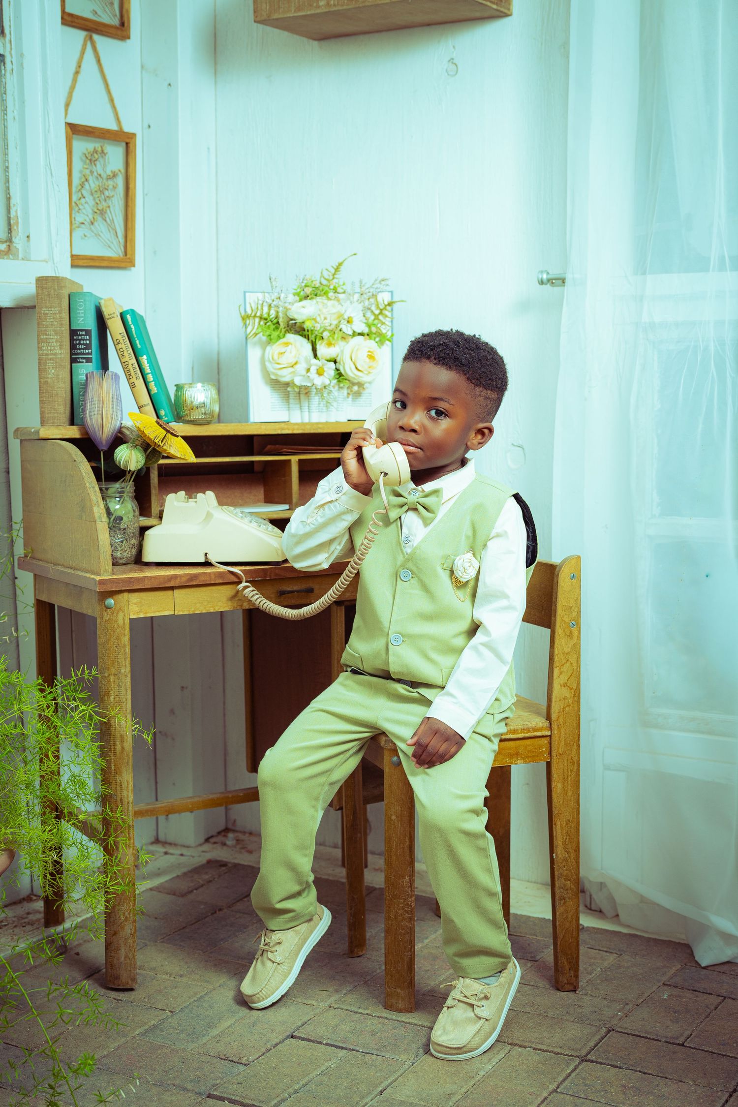 Young Black boy in mint green suit sits on chair holding vintage telephone receiver at antique wooden desk.