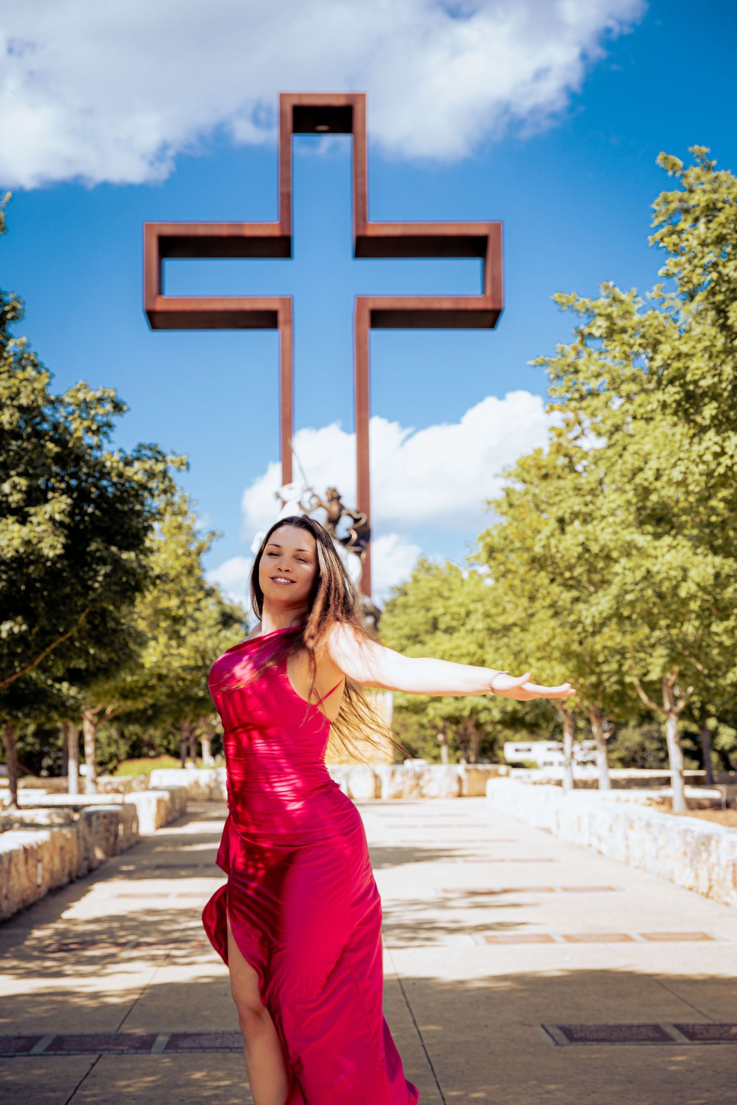 Woman in red dress posing joyfully in front of a large cross monument in a sunny park.