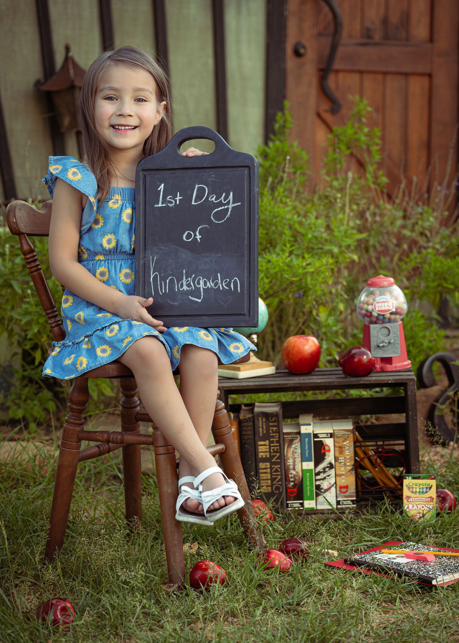 Young girl in blue dress sits on chair holding 1st Day of Kindergarten chalkboard sign, surrounded by apples and books.