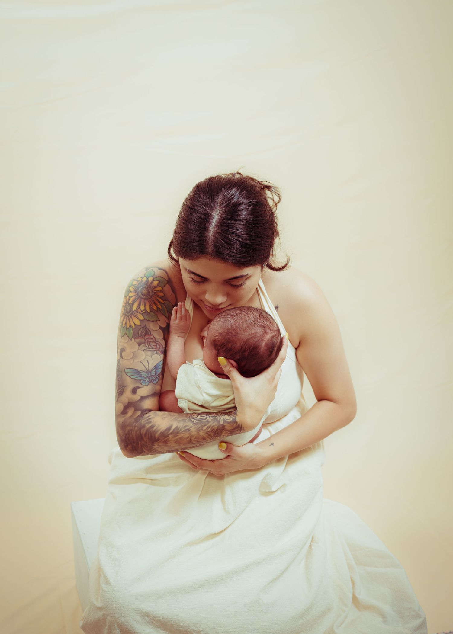 Tattooed mother in white dress tenderly cradling her newborn baby against a soft cream background.