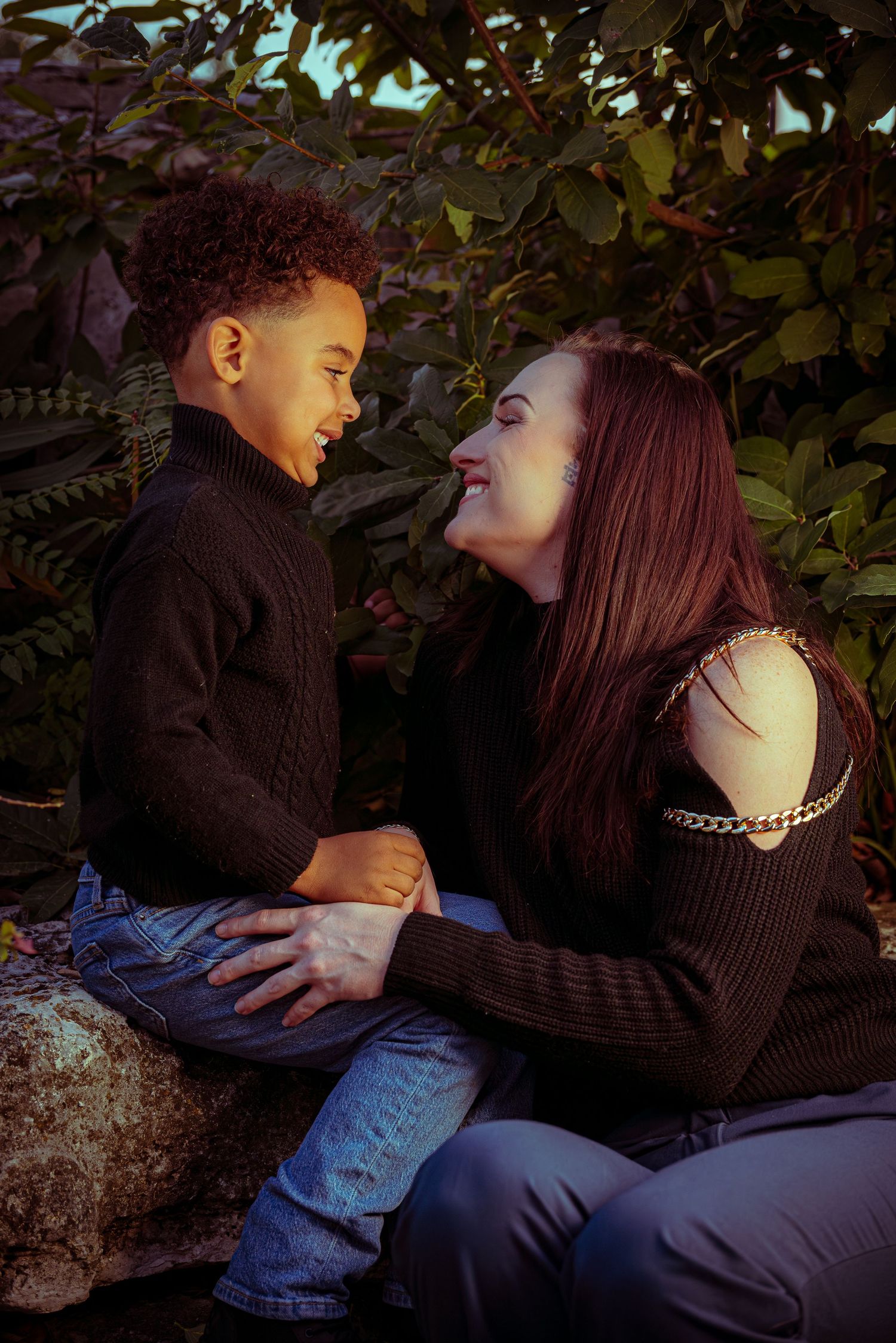 A couple in dark sweaters share an intimate moment while sitting together on a rock in a garden setting.