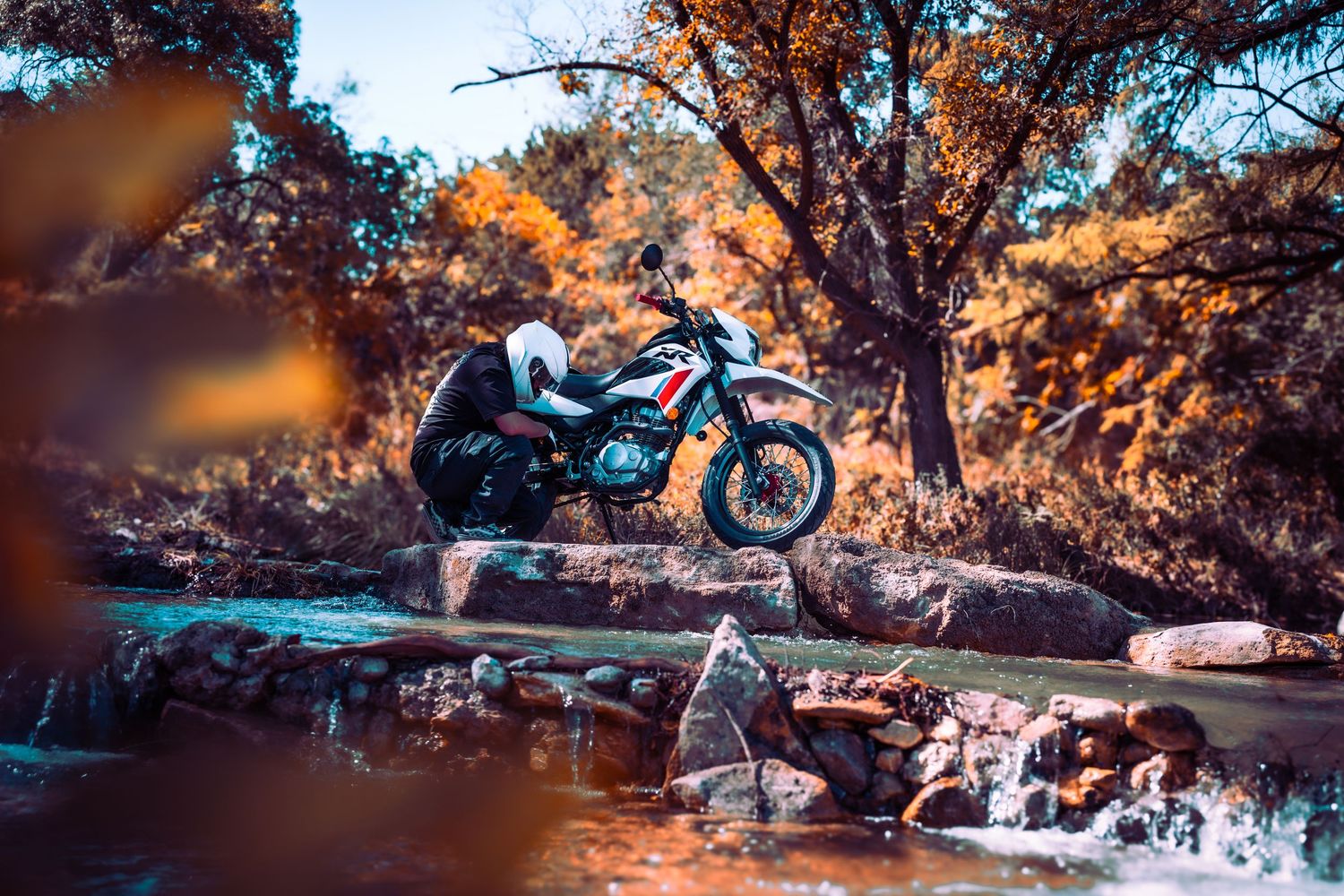 Adventure motorcycle staged near flowing stream with sunlit fall trees in background.