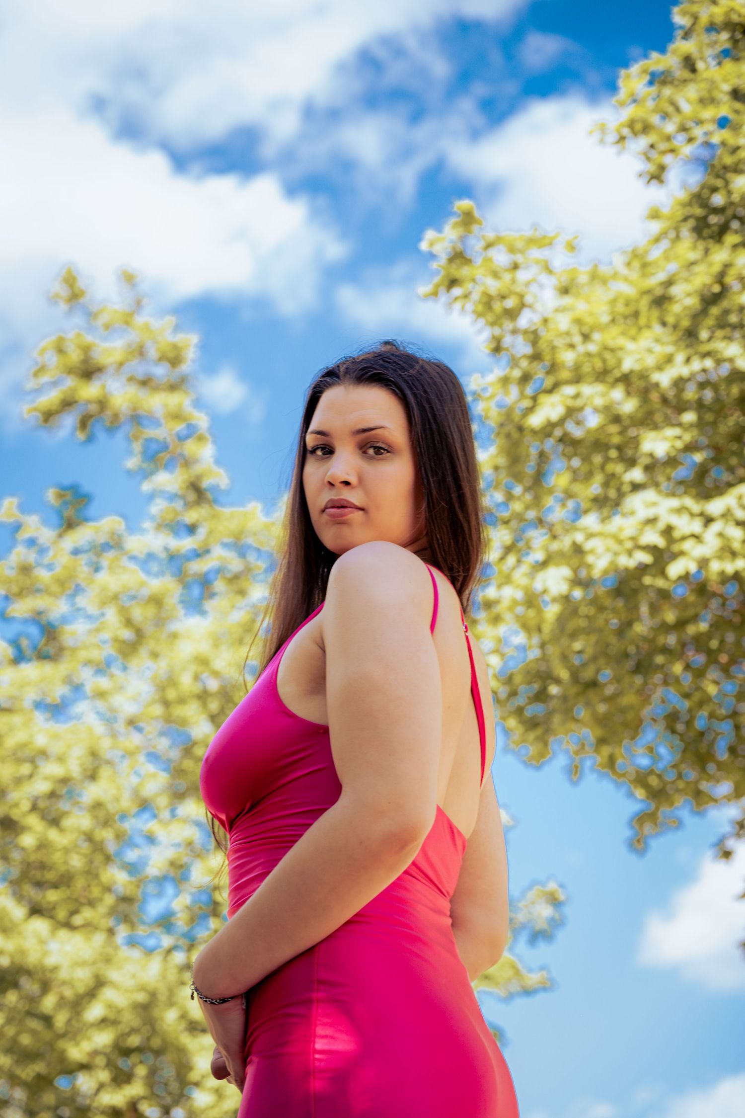 Young woman in pink dress poses outdoors with green trees and blue sky background.