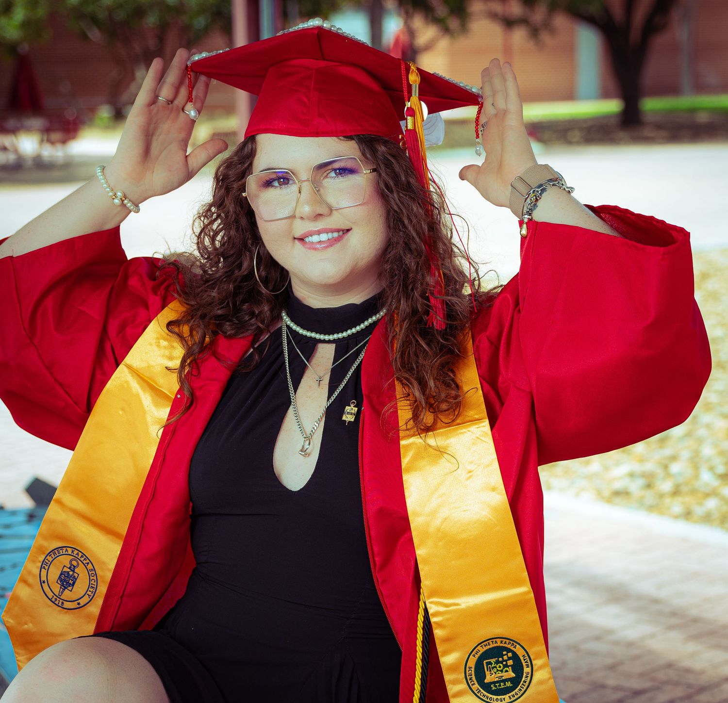 Smiling graduate in red and gold cap and gown poses outdoors holding mortarboard on college campus.
