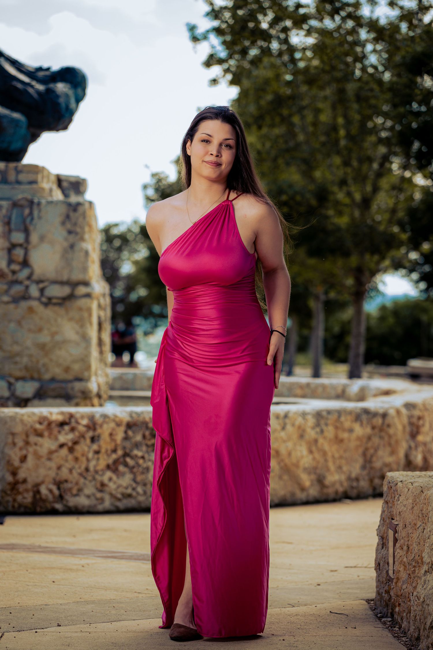 Woman in elegant pink one-shoulder gown posing outdoors near stone fountain and trees.
