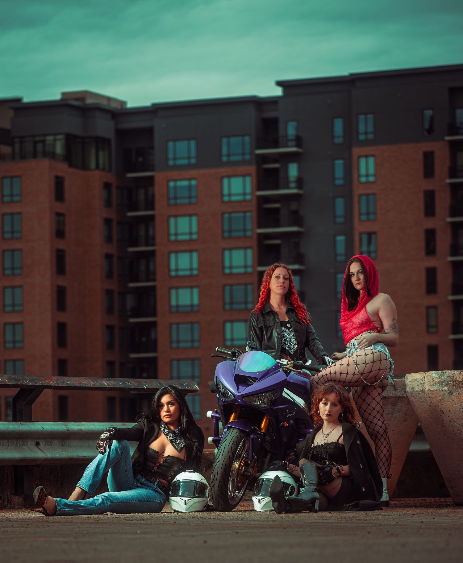 Four people lounging on a rooftop with backpacks and belongings against an urban backdrop of brick apartment buildings.