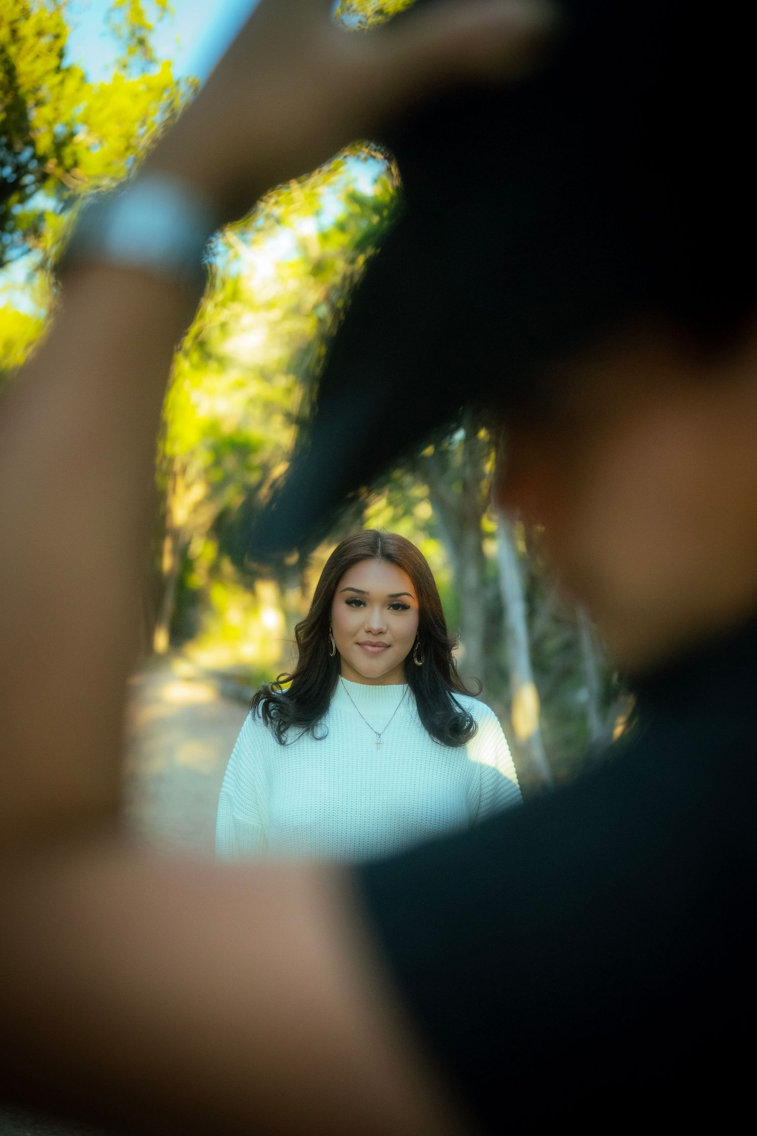 Woman in white top smiles softly on a tree-lined path, framed by a blurred foreground figure.