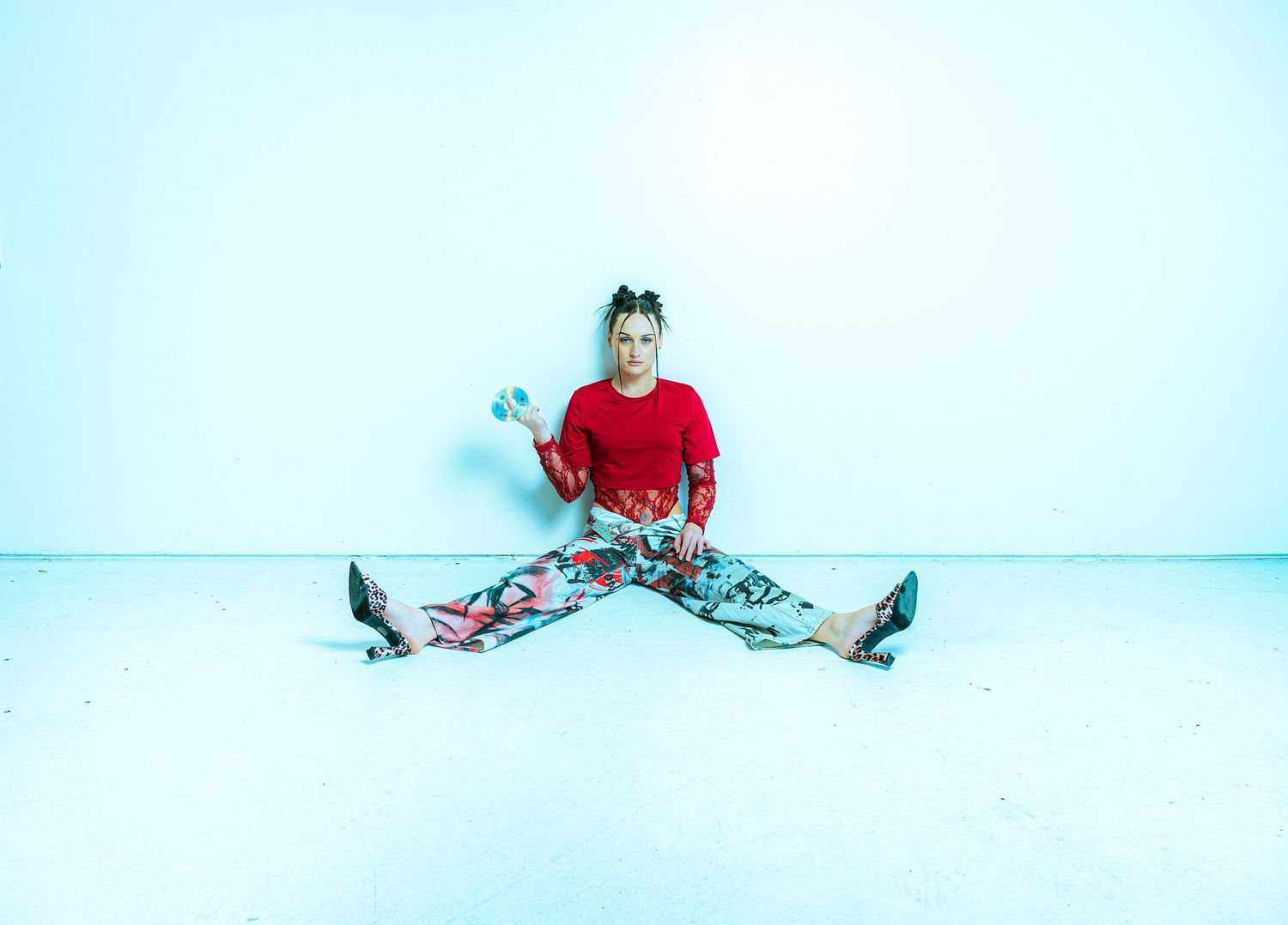 Woman in red top and colorful pants doing splits against white wall, holding blue accessories.