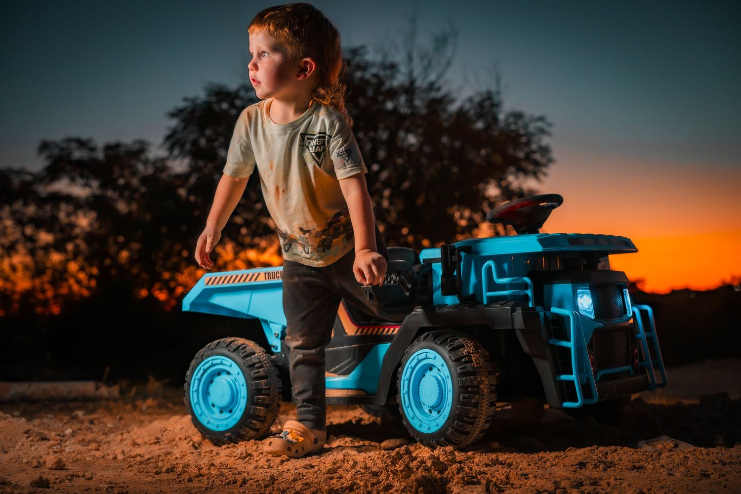 Child riding blue toy tractor at sunset with dramatic lighting.