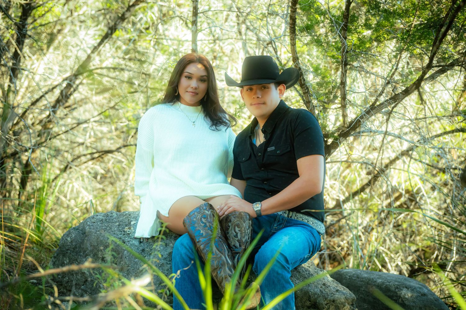 A couple sits together in a sunlit forest setting surrounded by lush green foliage and dappled sunlight.