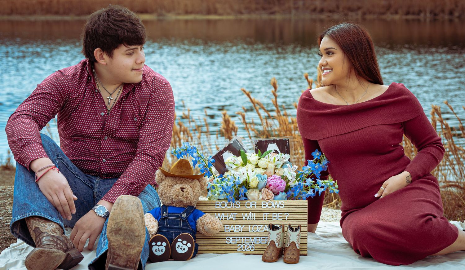 Couple sitting outdoors near a lake with teddy bears and flowers between them during a maternity photoshoot.