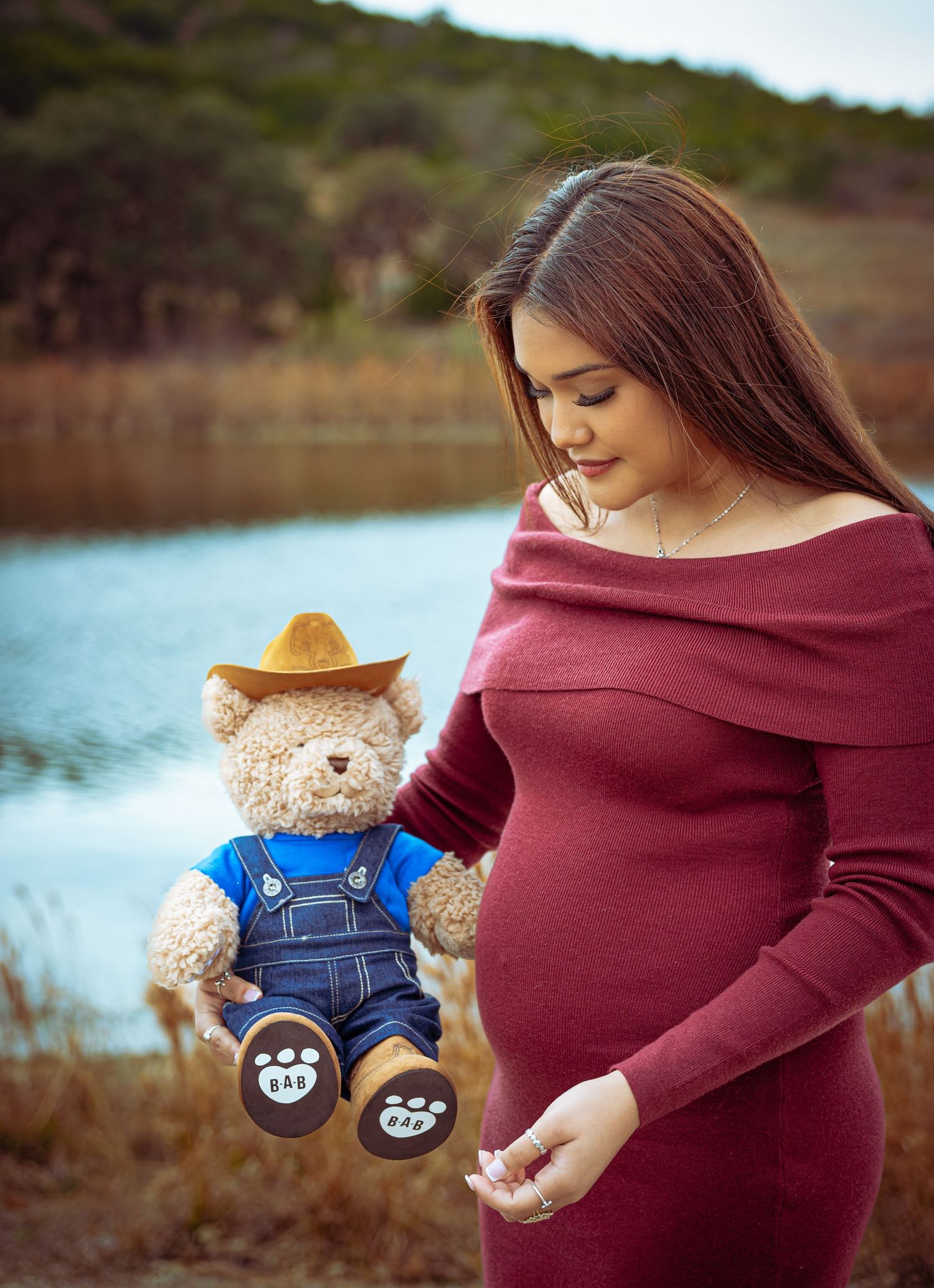 Pregnant woman in red off-shoulder dress holds teddy bear in cowboy hat near lake during maternity photoshoot.