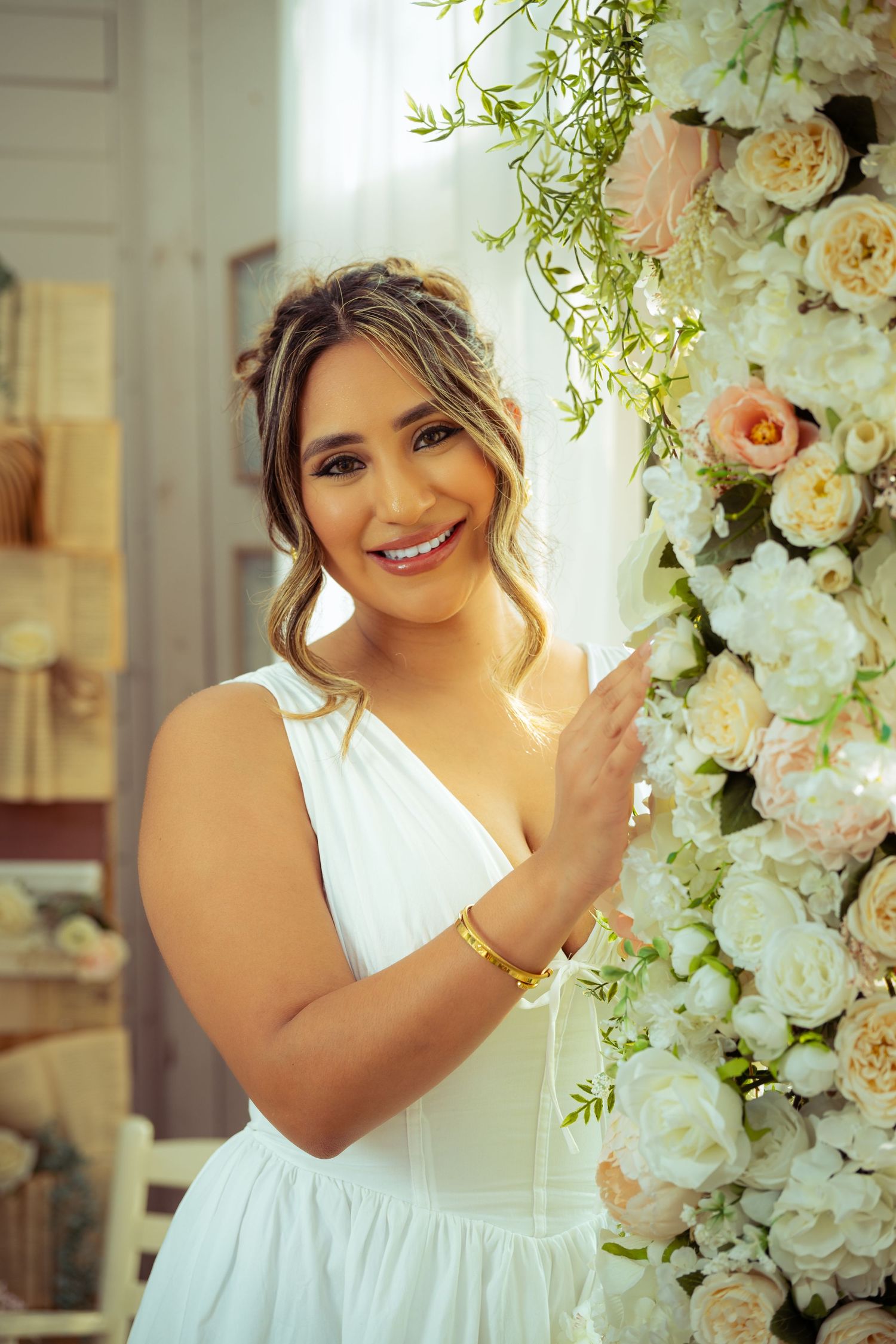 Smiling woman in white dress holding white and pink floral arrangement, wearing gold bracelet in bright elegant setting.