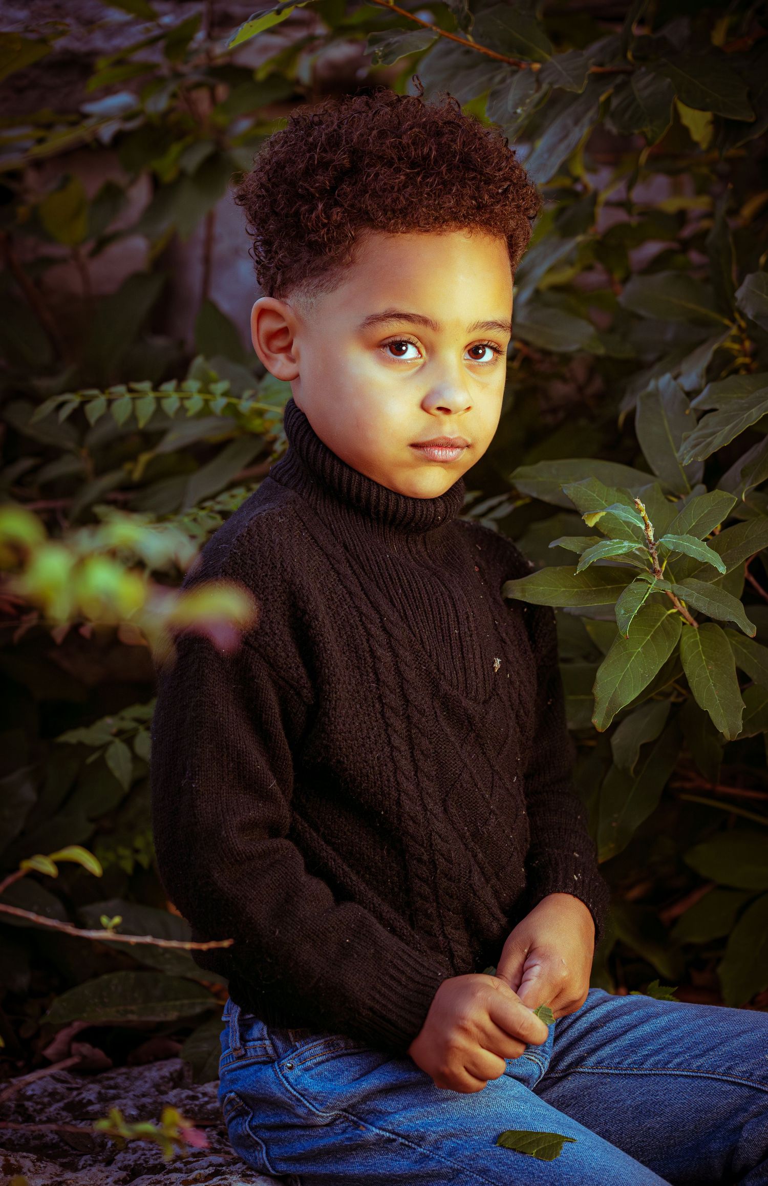 A young child in a dark knit sweater sits outdoors against a backdrop of green foliage.