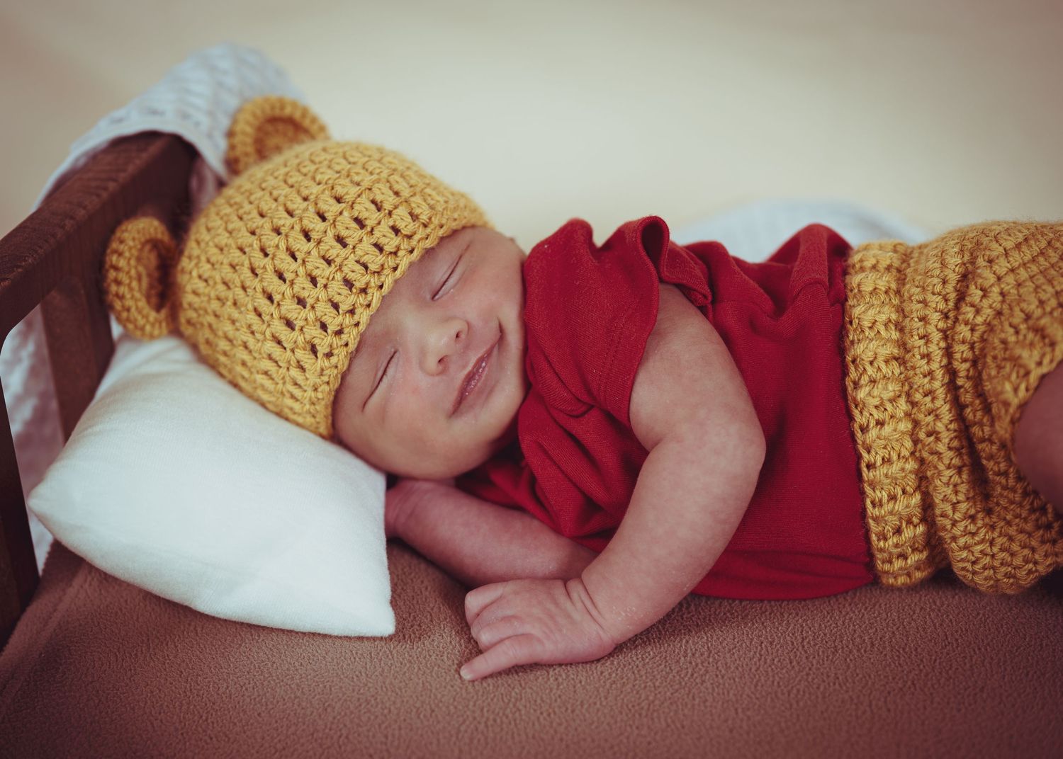 Close-up of sleeping newborn in red outfit and yellow crocheted bear ear hat on white pillow.