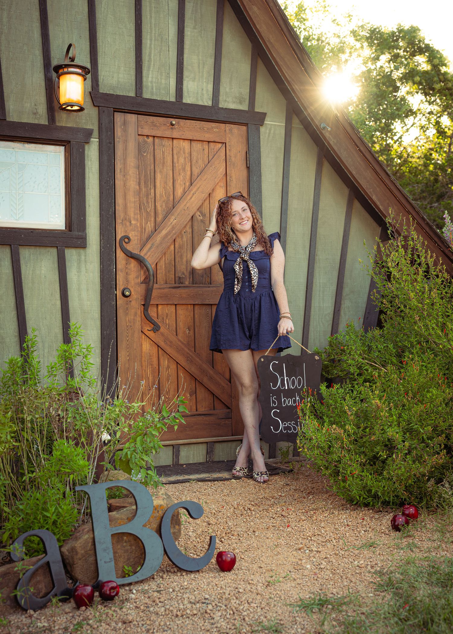 Young woman in blue dress poses by rustic green cottage door with ABC letters and apples for back-to-school photo session.