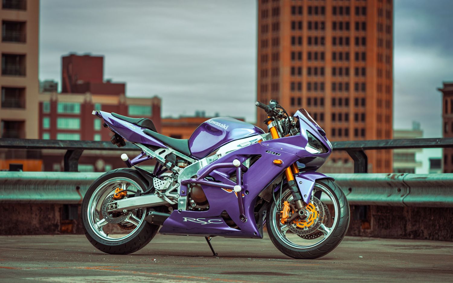 Four purple and silver sports motorcycles parked in a row against an urban backdrop with brick buildings.