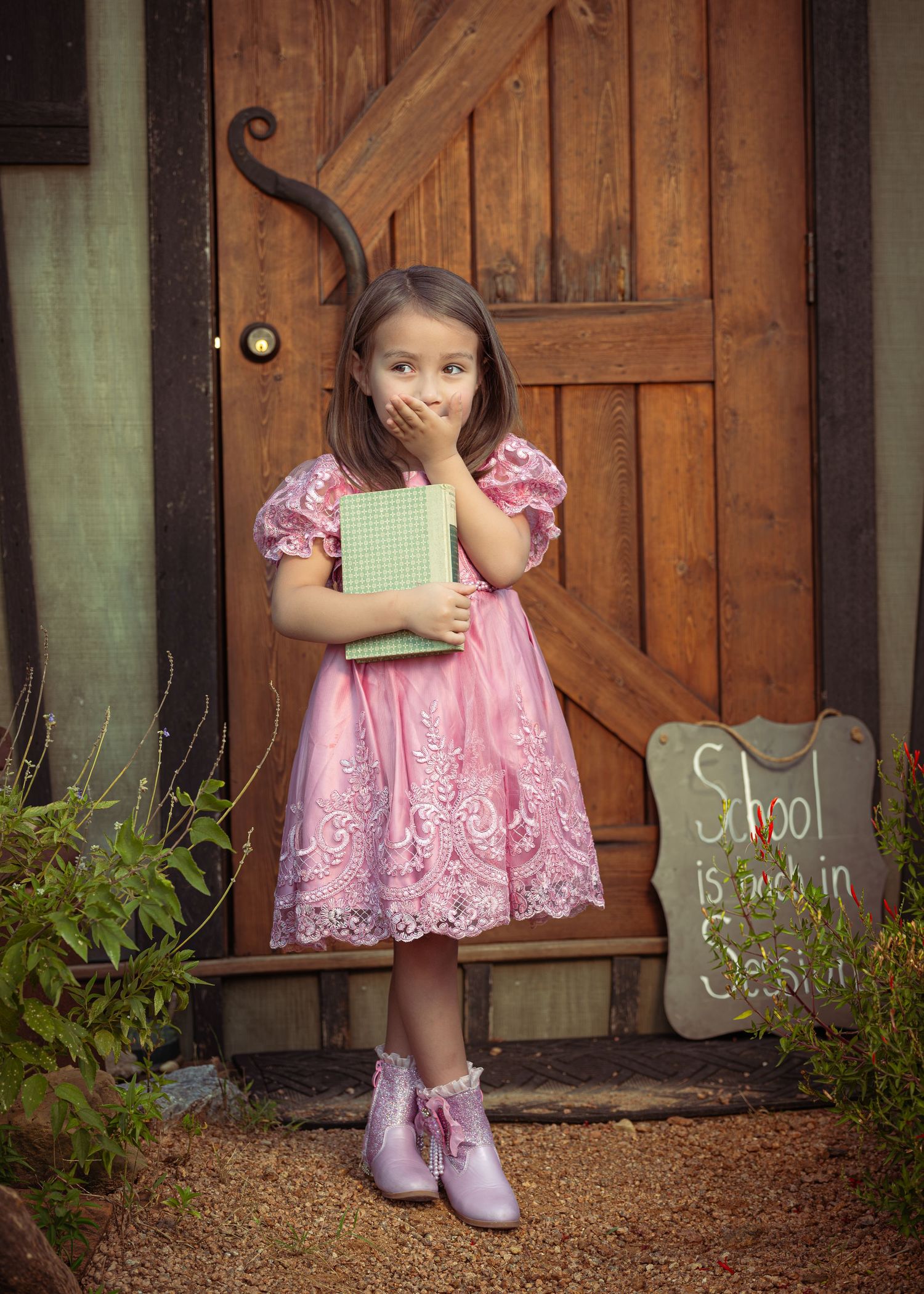 A young girl in a pink lace dress holds a green book by a wooden door with a School is in Session chalkboard sign.