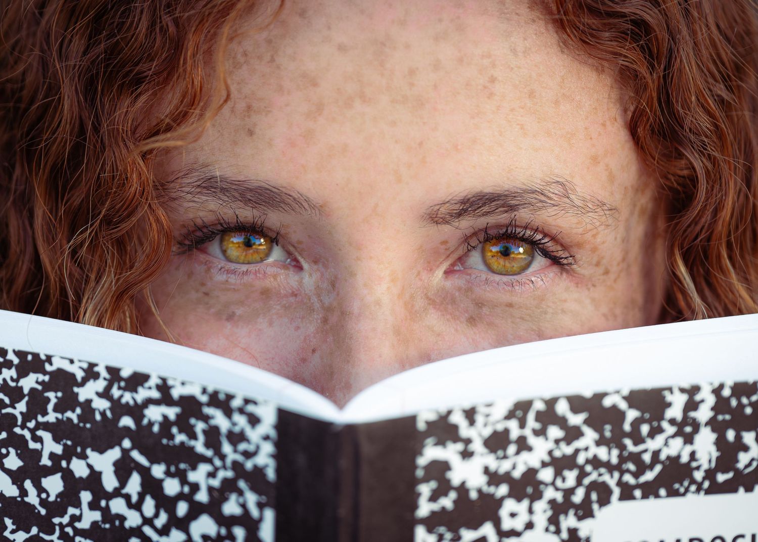 Close-up of a freckled redhead woman with striking amber eyes peering over an open composition notebook.