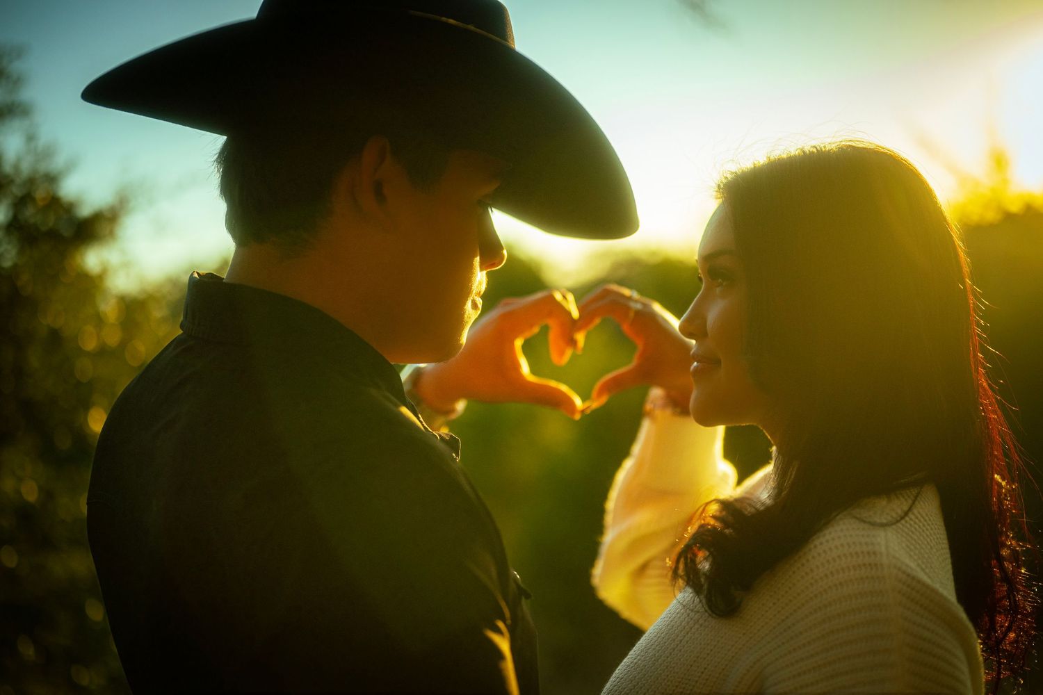 Couple making heart shape with hands at golden sunset, man in cowboy hat, romantic outdoor portrait.