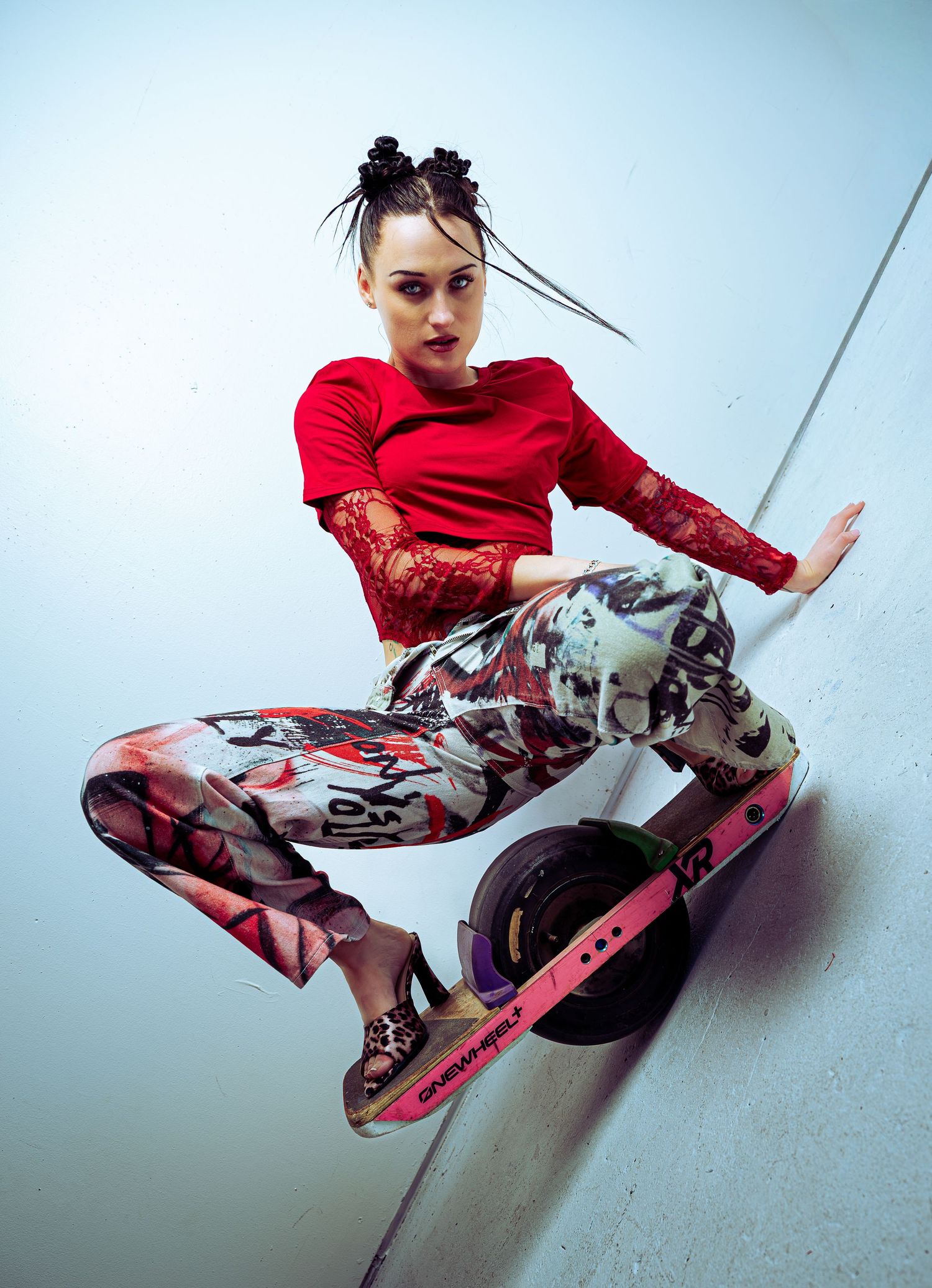 Woman in red lace top and graffiti pants balancing on an electric scooter in a dynamic pose.