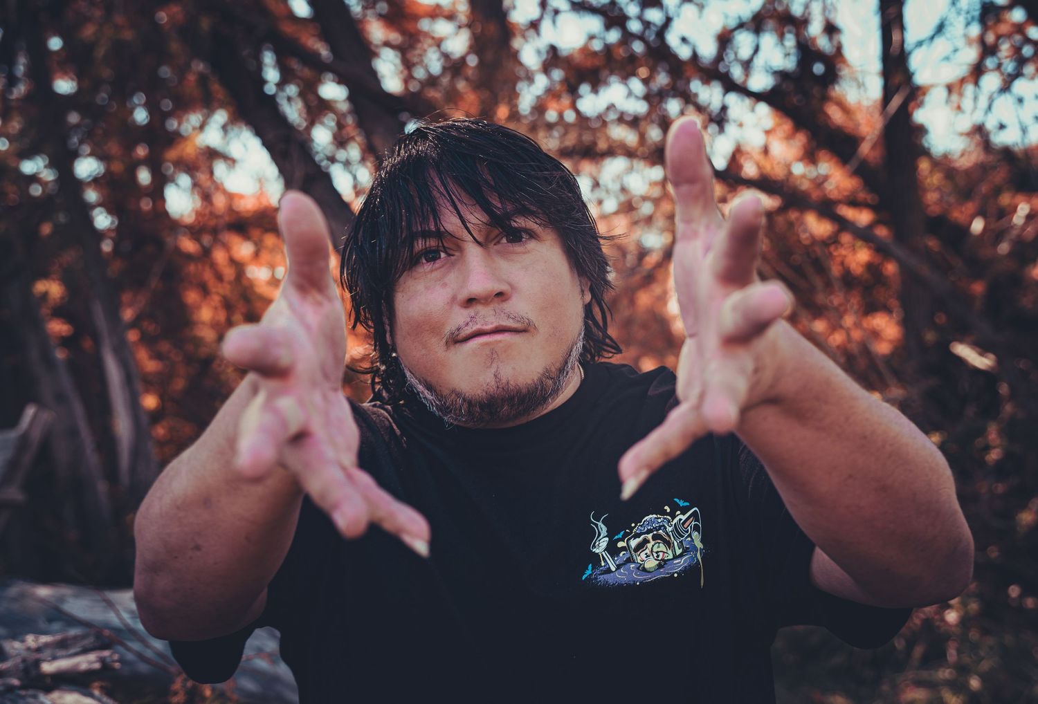 Person making hand gestures while seated outdoors with autumn foliage in the background.