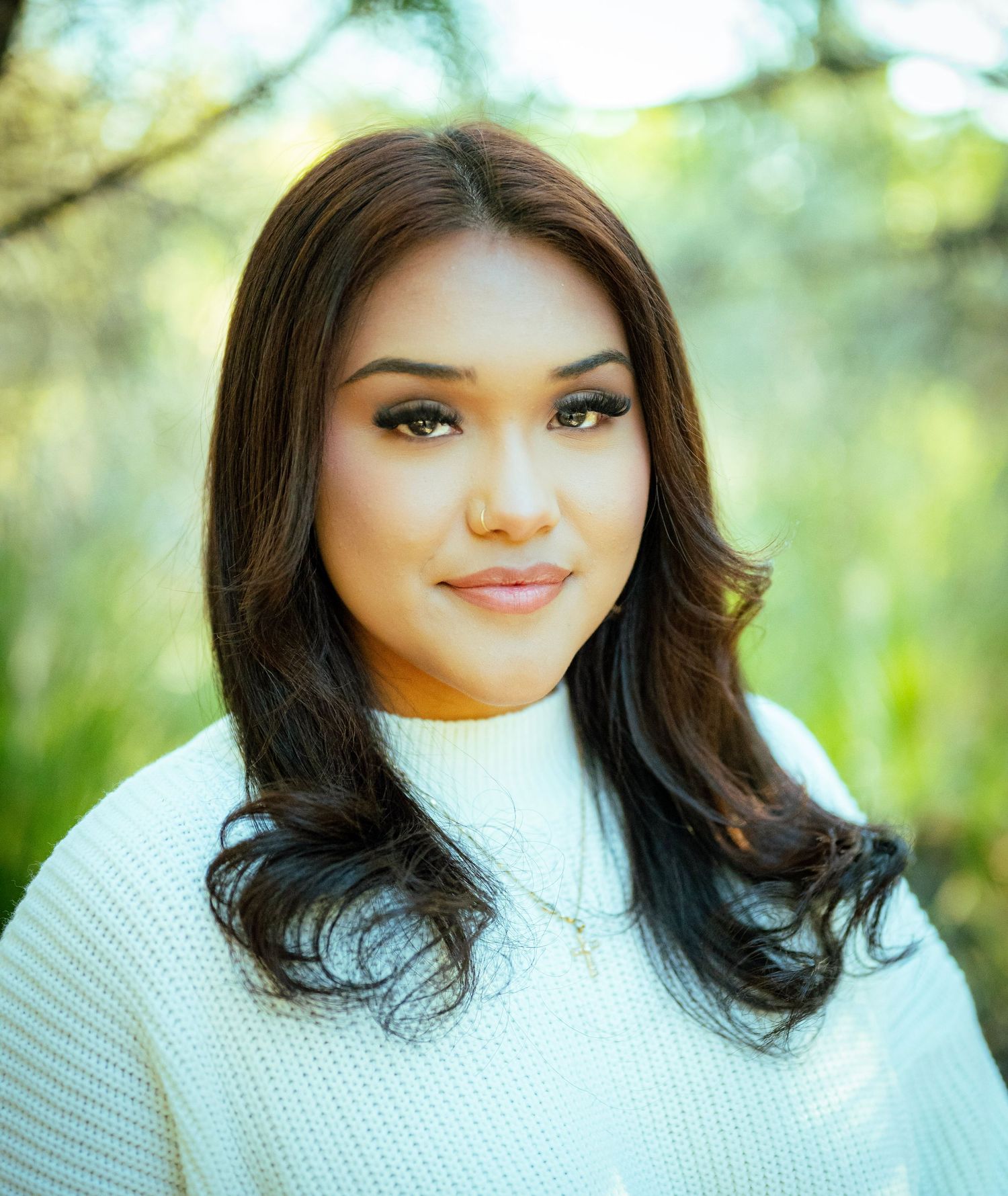 Portrait photo outdoors with soft natural lighting and white sweater against blurred green foliage background.