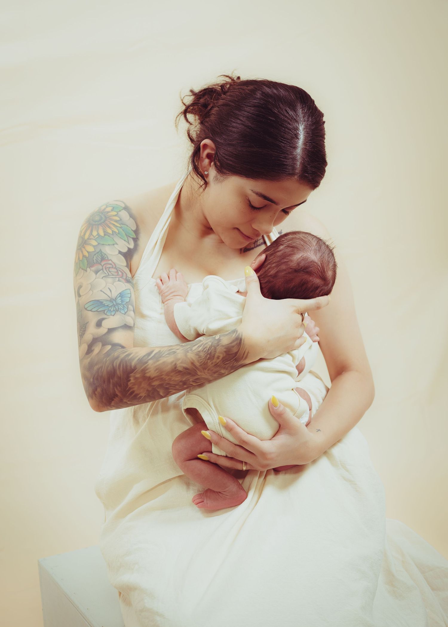 Tattooed mother tenderly holding and kissing her newborn baby, dressed in white, against a soft background.