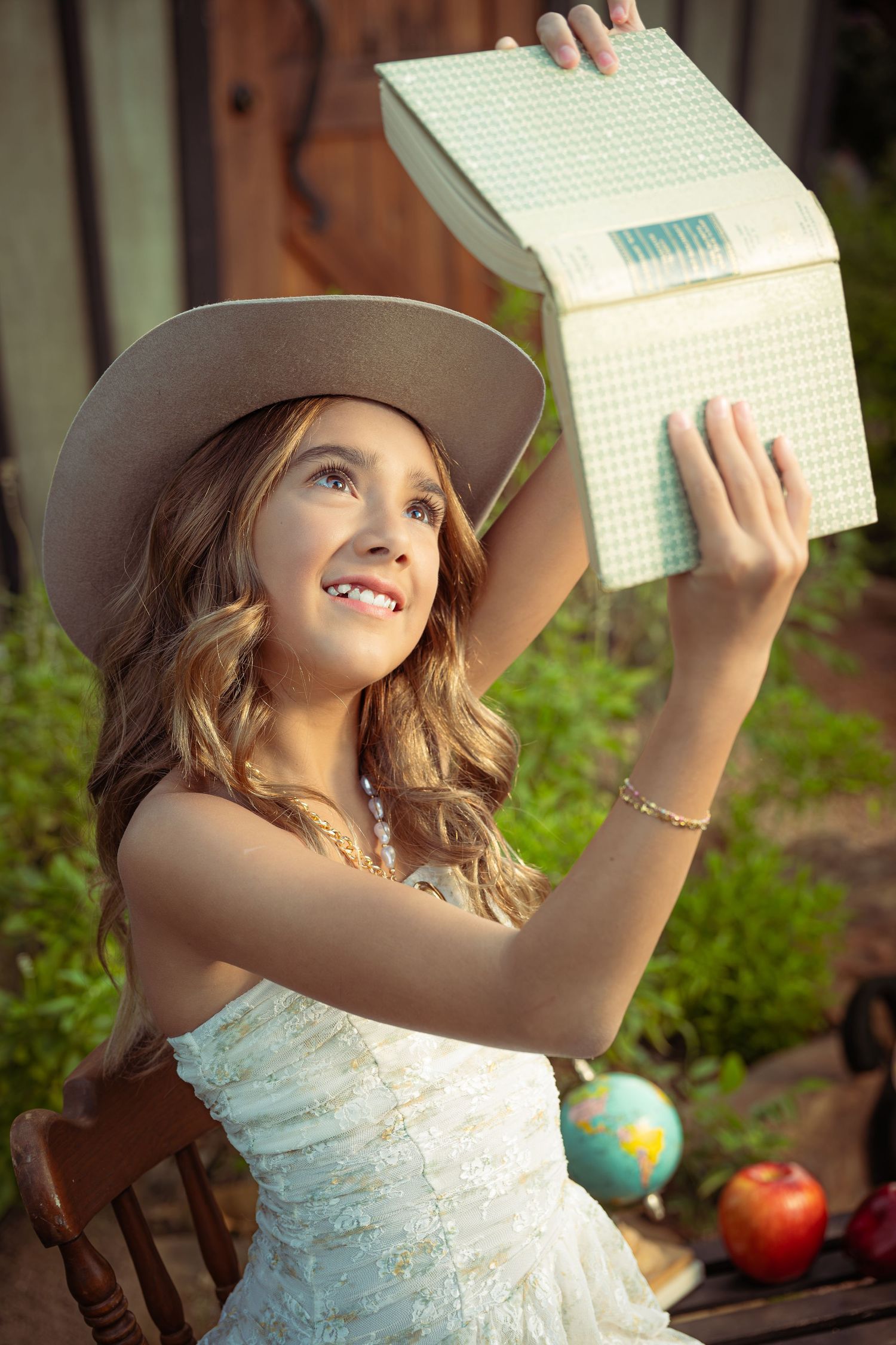 A smiling young girl in a white dress and hat holds up a green gift box outdoors near a globe and apple.