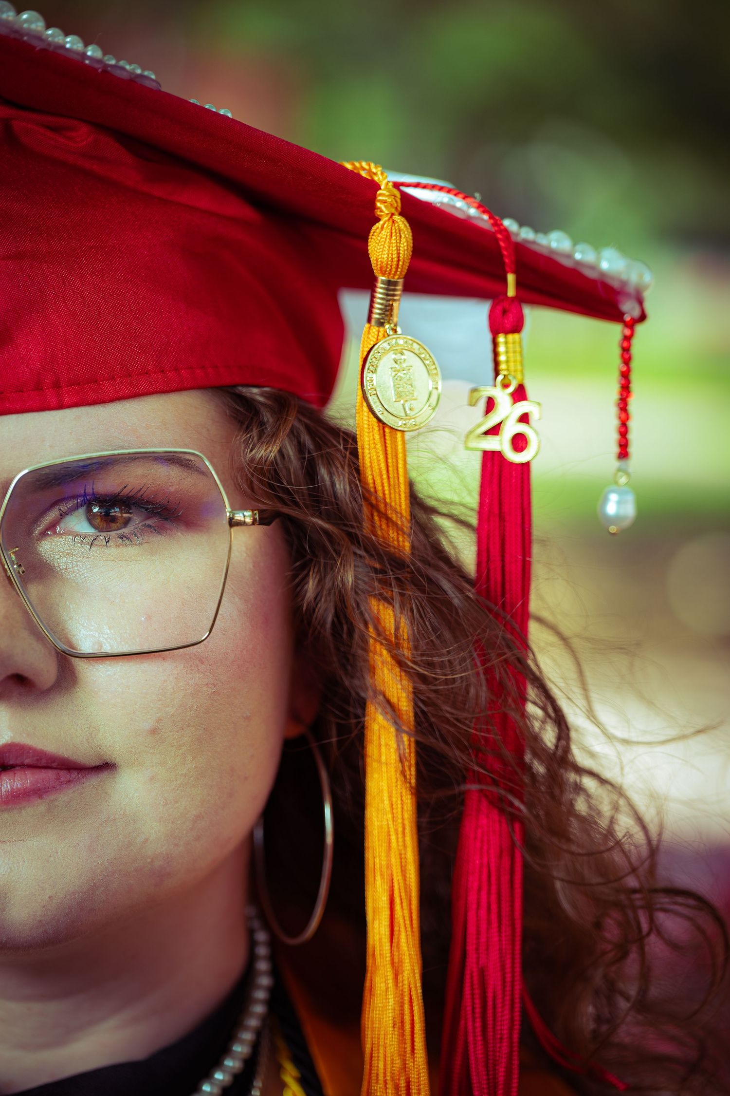 Close-up of a young woman in a red graduation cap with gold tassel and 2026 charm, wearing pearl jewelry and glasses.