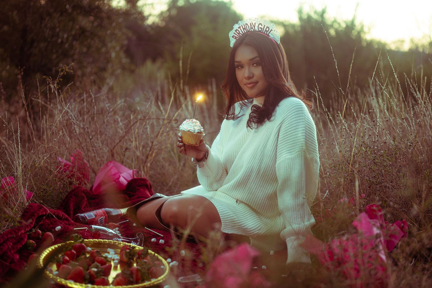 A person in a mint green sweater sits outdoors at sunset among pink flowers in a dreamy garden setting.