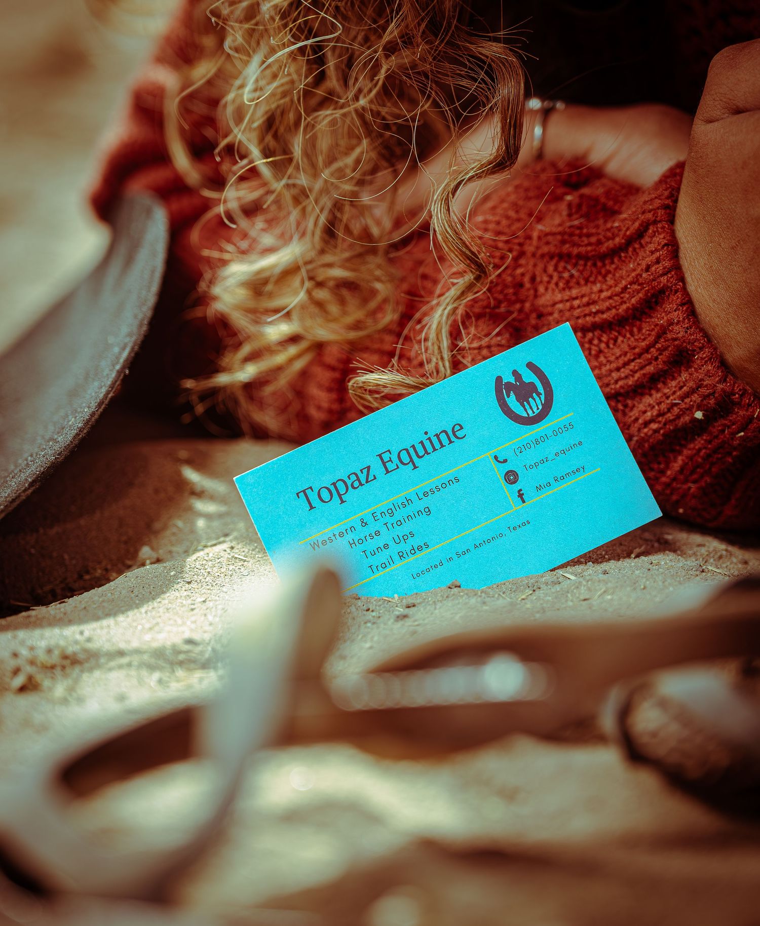 A bright turquoise business card for Topaz Equine rests on a surface next to a red knit sweater and blonde curls.