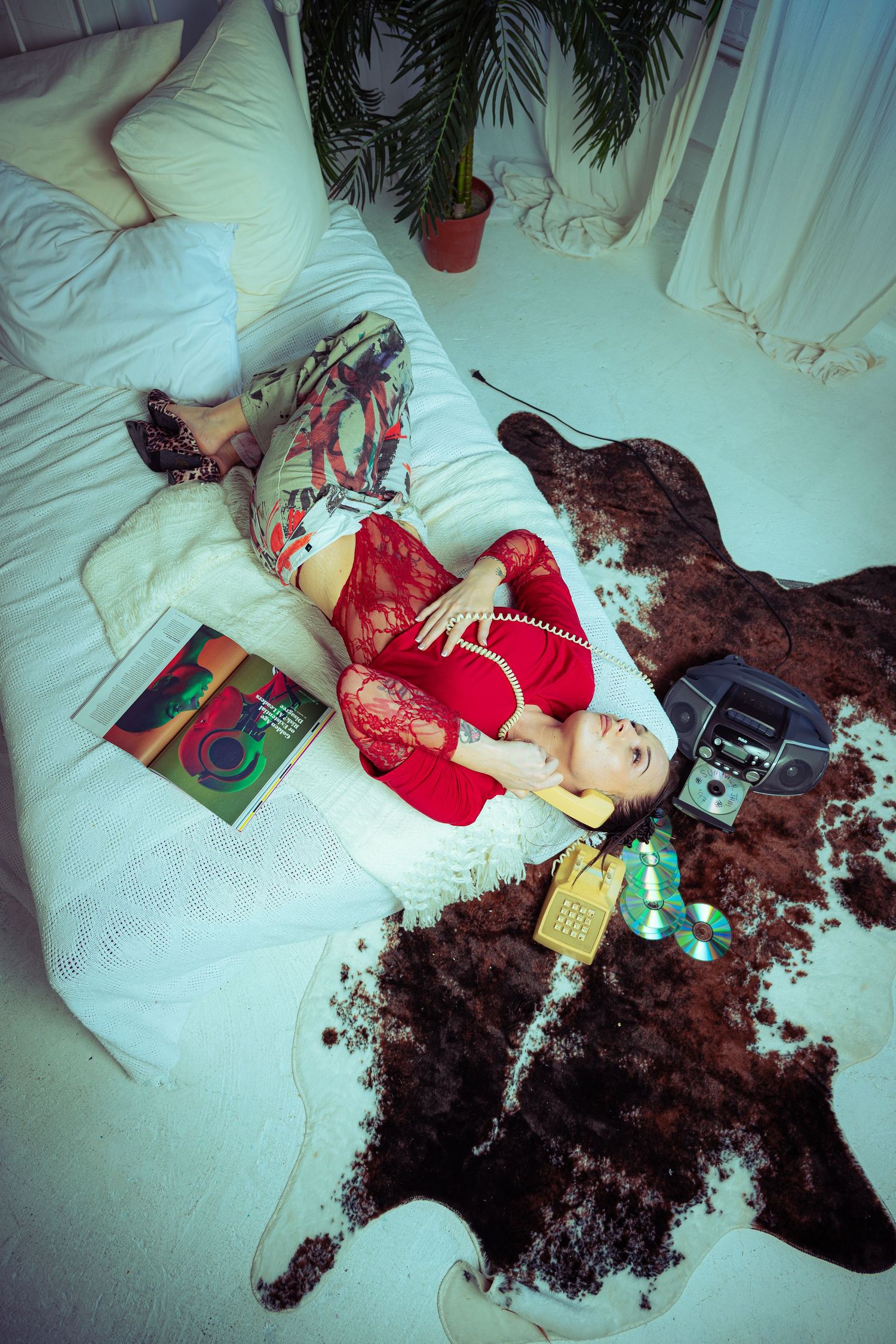 Woman in red lace top lying on bed with vintage phone, boombox, CDs, and colorful magazine on cowhide rug.
