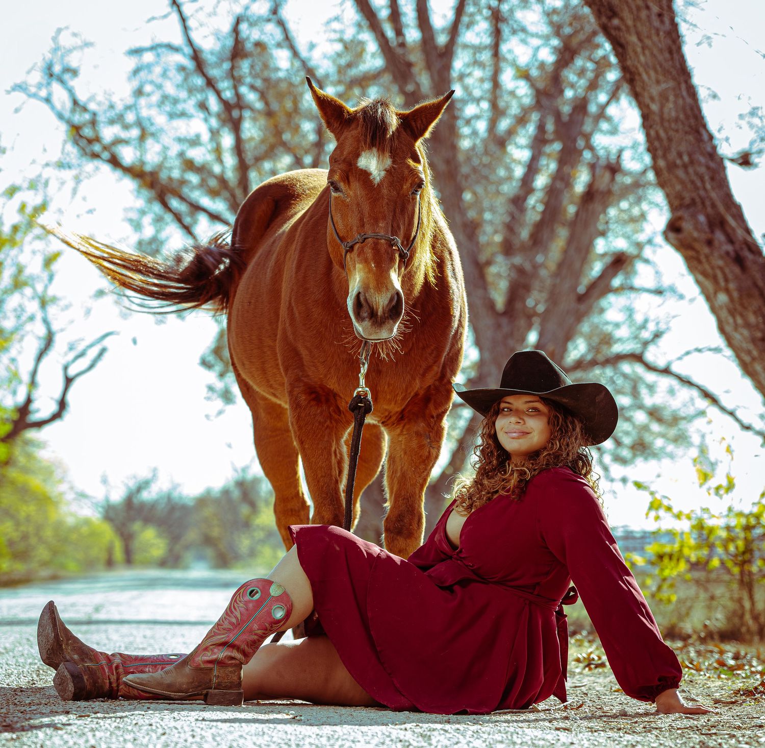 A chestnut horse stands beside someone in a red dress and cowboy boots sitting on the ground by trees.