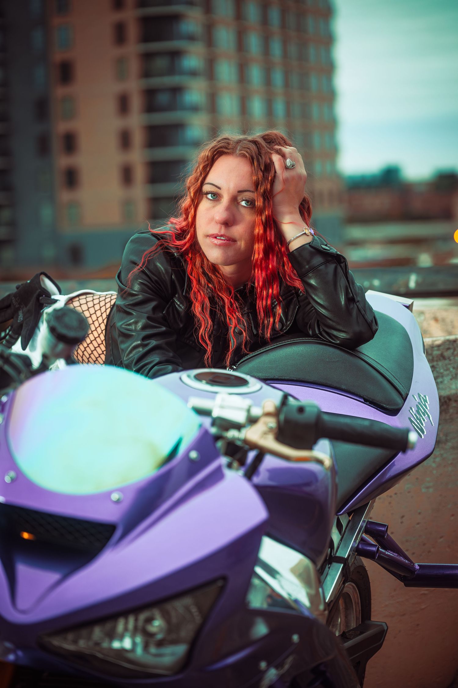 A person with vibrant red hair poses with a purple motorcycle against an urban backdrop.